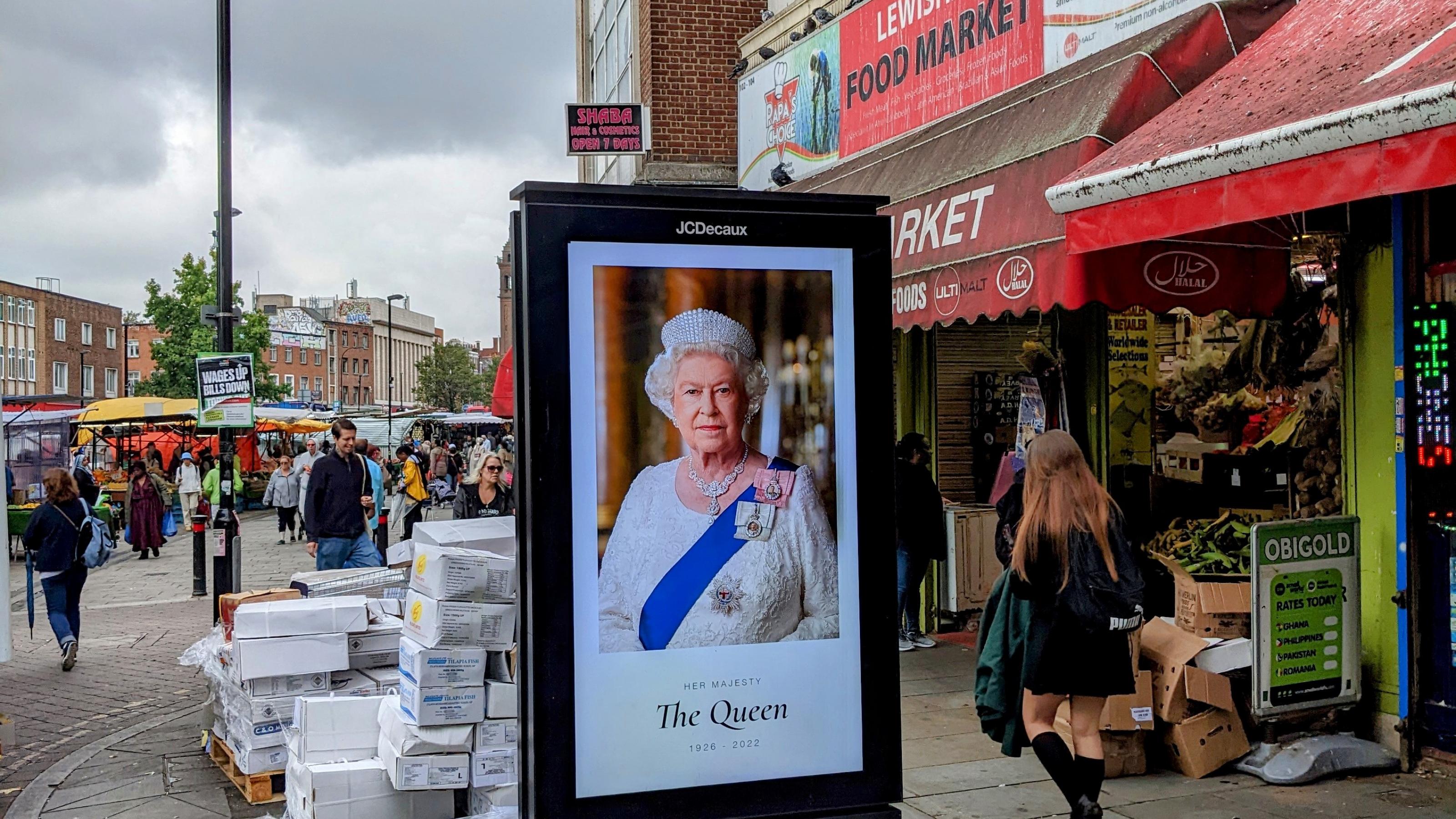 Ein Leuchtplakat mit dem Bild der Queen steht in einem Straßenmarkt.