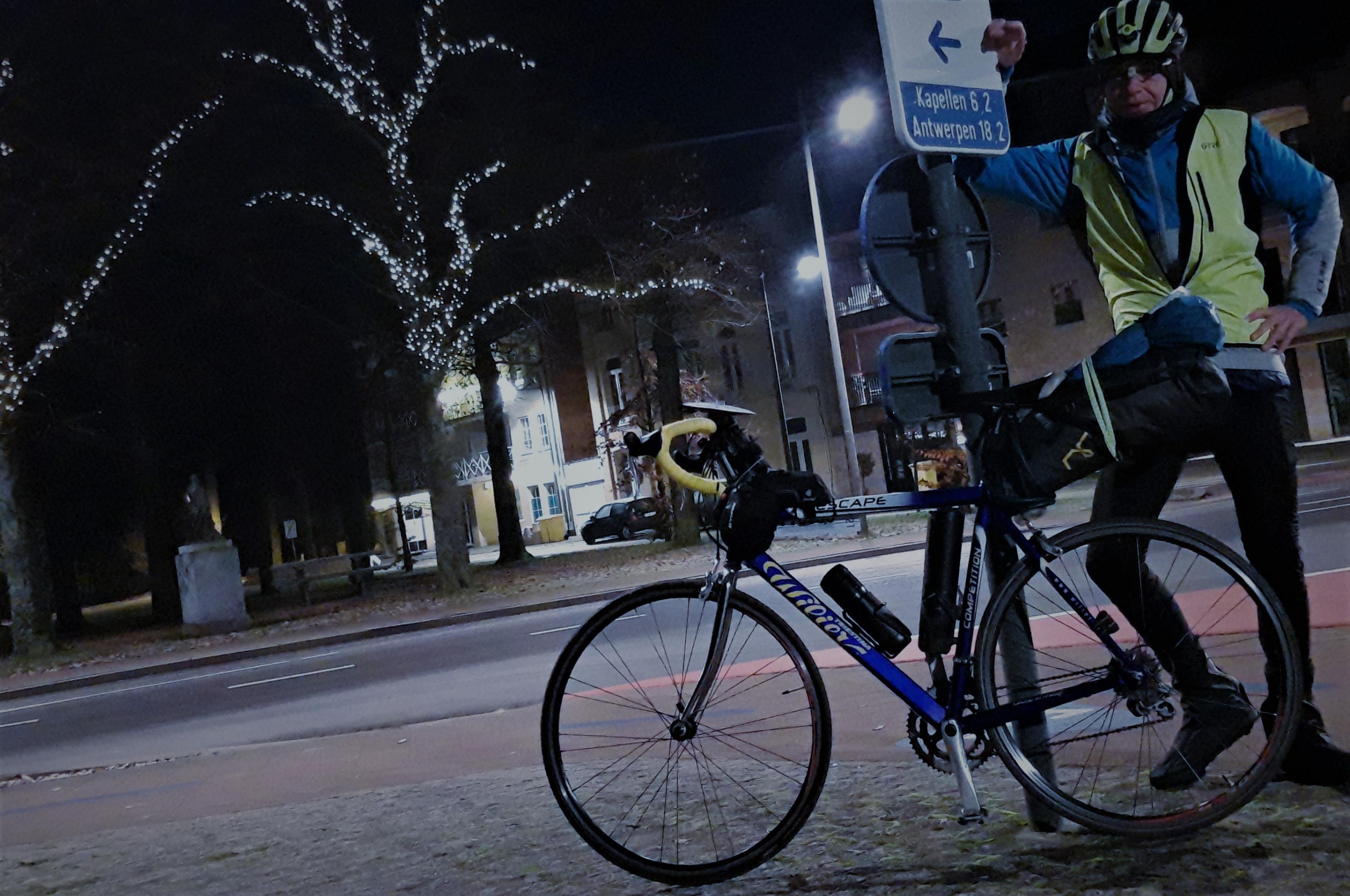 Der Autor lehnt, in Radkluft und in weihnachtlich-näöchtlicher Szenerie, an einem Wegweiser, auf dem vermerkt ist „Antwerpen 18,2 km“.