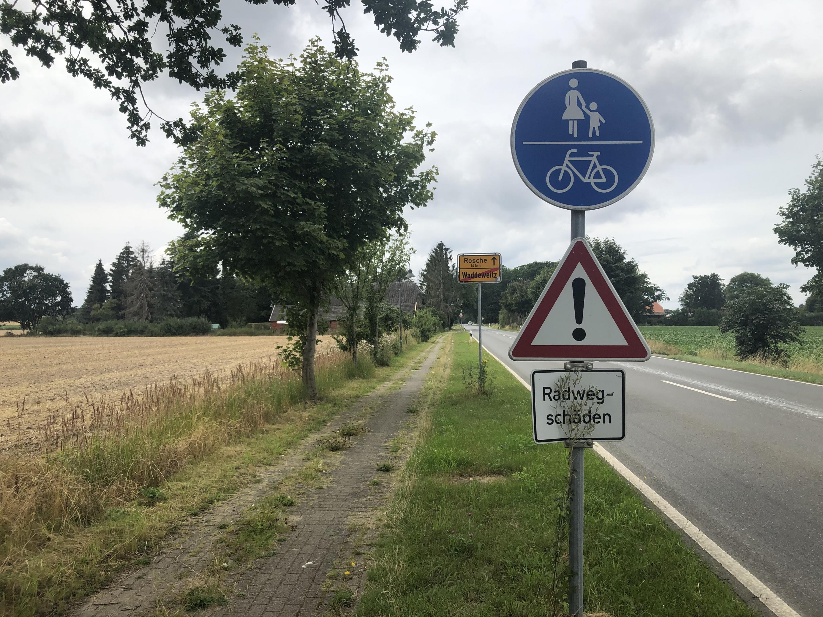 Das Bild zeigt einen Radweg an einem Ortsrand in Niedersachsen, der als benutzungspflichtig ausgeschildert ist, aber in einem sehr schlechten Zustand ist. Darauf weist zugleich ein Schild hin, das vor Radwegschäden warnt.