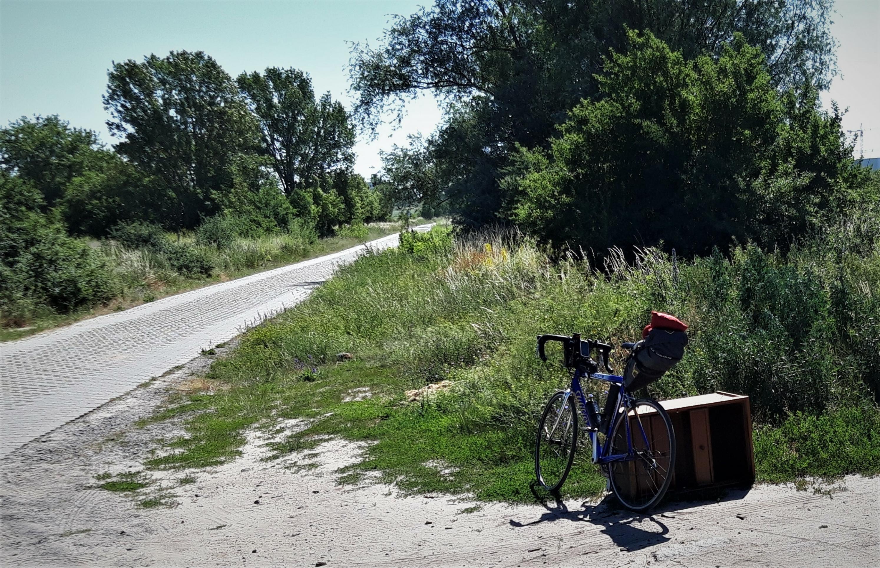 Links ein Kolonnenweg, umrandet von Gebüsch; rechts das Fahrrad, das an einem kaputten, illegal abgelagertem Holzmöbel lehnt.