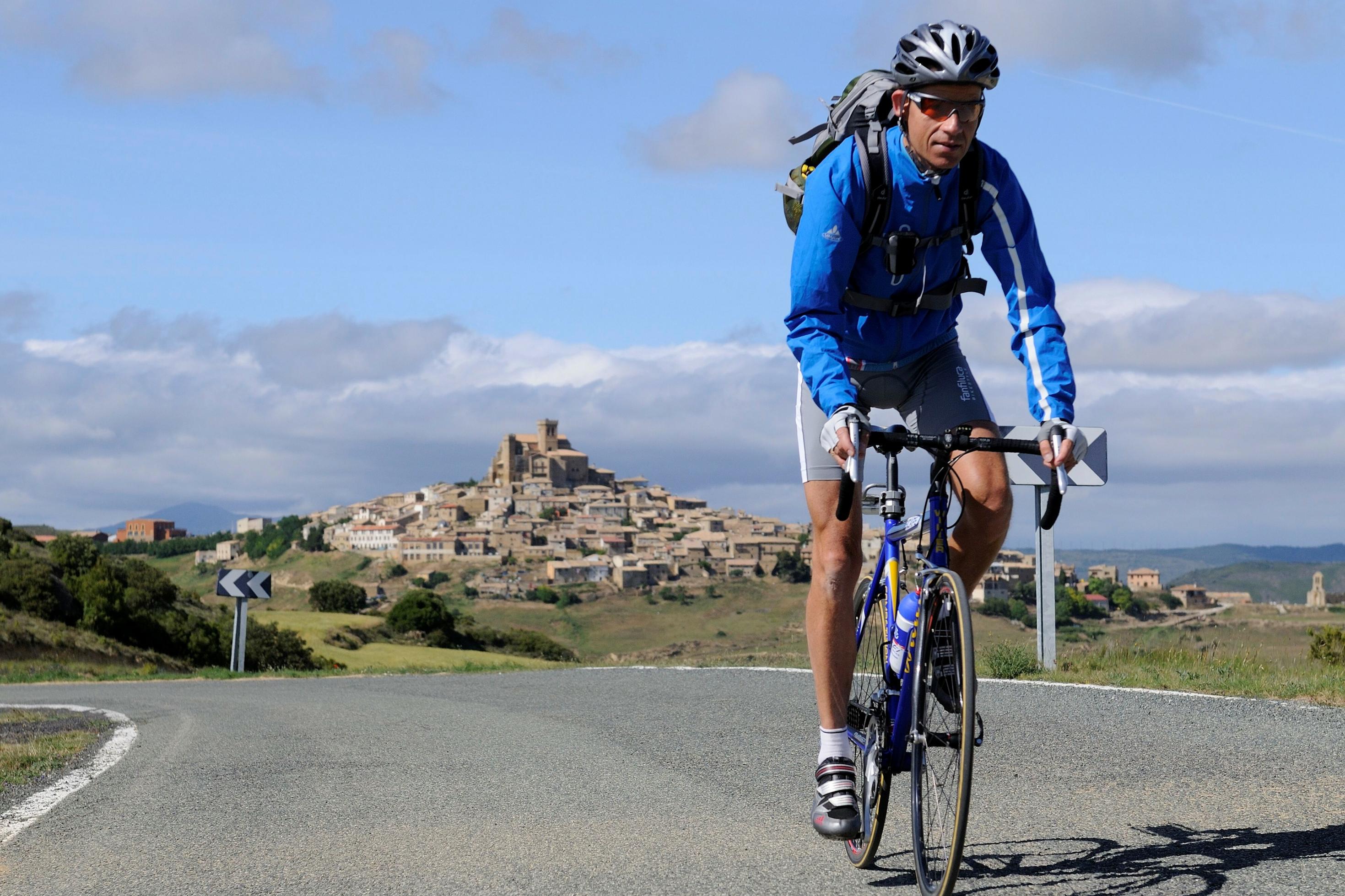 Gebirgsszene mit Mittelalter-Dorf im Hintergrund; im Vordergrund eine kurvige Landstraße, auf der der RadelndeReporter per Rennrad ins Bild fährt.