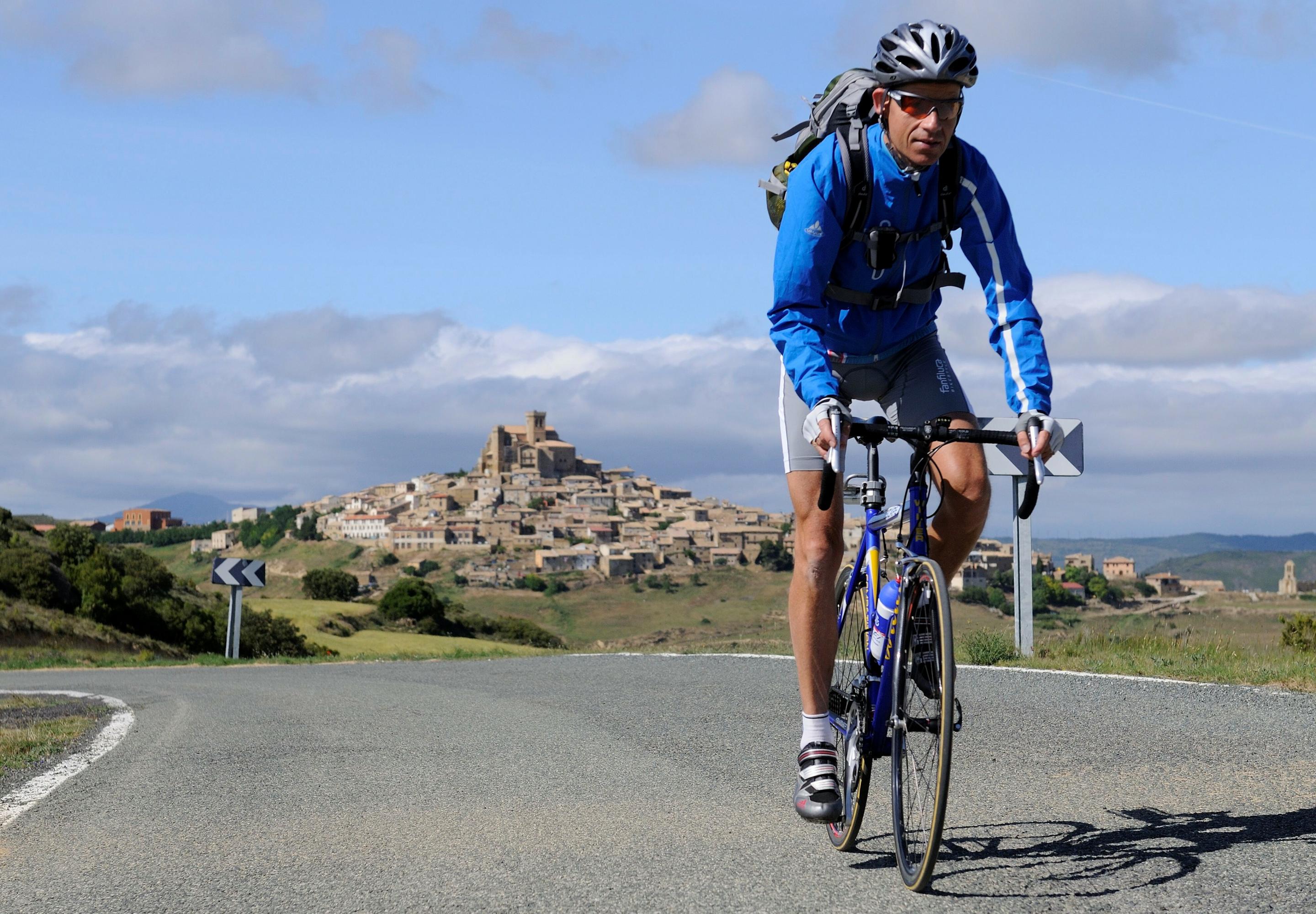 Gebirgsszene mit Mittelalter-Dorf im Hintergrund; im Vordergrund eine kurvige Landstraße, auf der der RadelndeReporter per Rennrad ins Bild fährt.