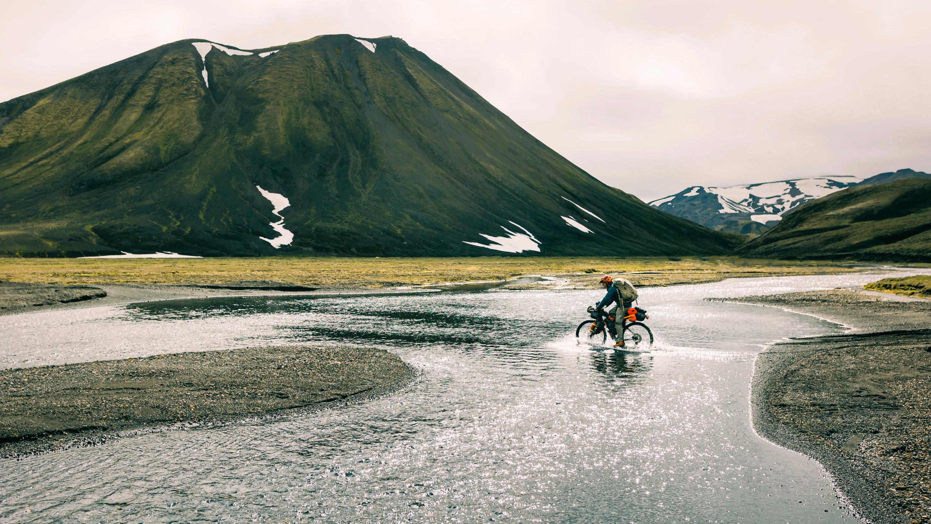 Ein in wilder isländischer Landschaft einsamer Radfahrer quert einen breiten Wasserstrom, der ihm beim „Pedalieren“ nasse Füße bereitet.