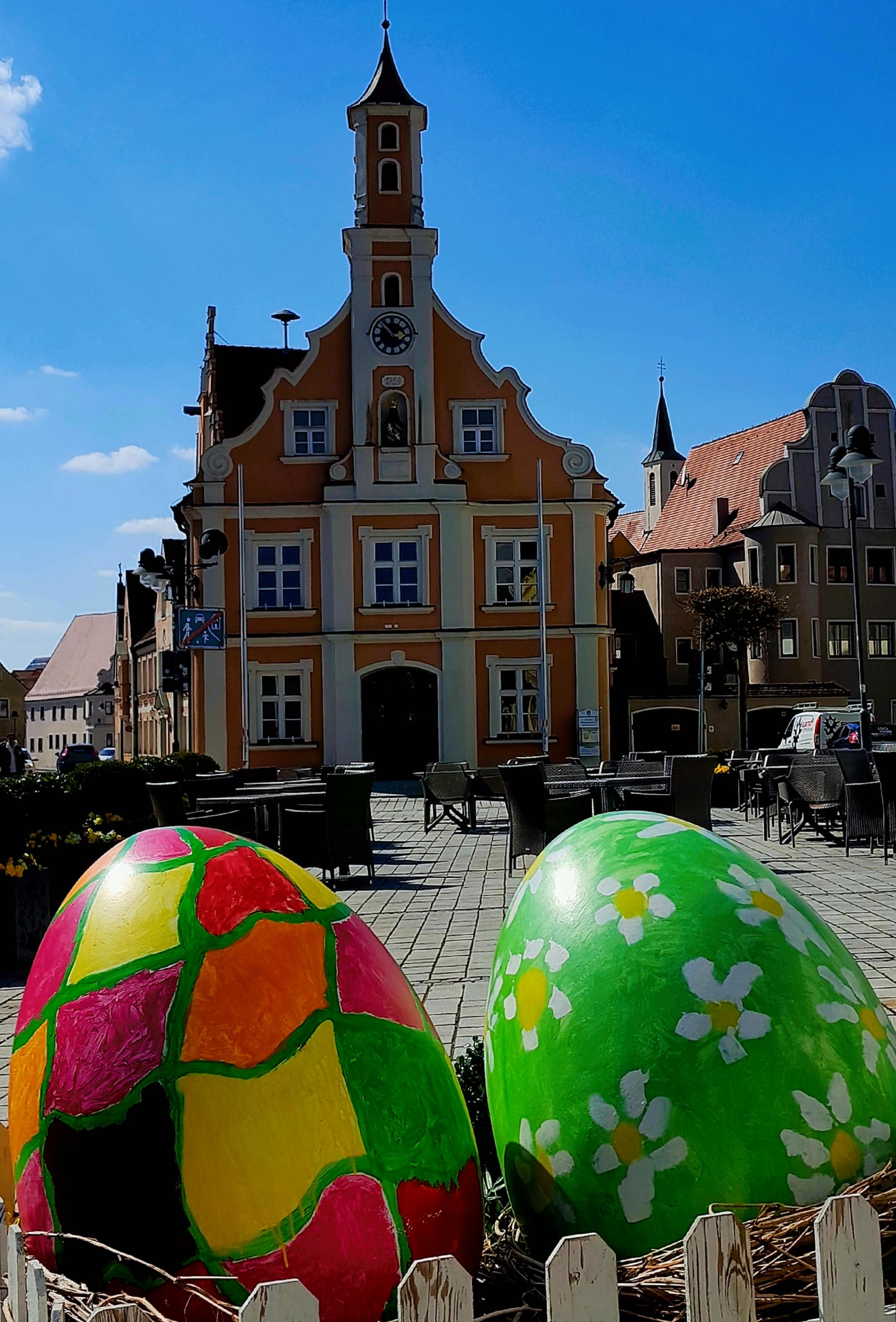 Im Vordergrund zwei sonnenbeschienene, quietschbunt bemalte Eier; im Hintergrund ein Bayerisch-schwäbisches Rathaus.