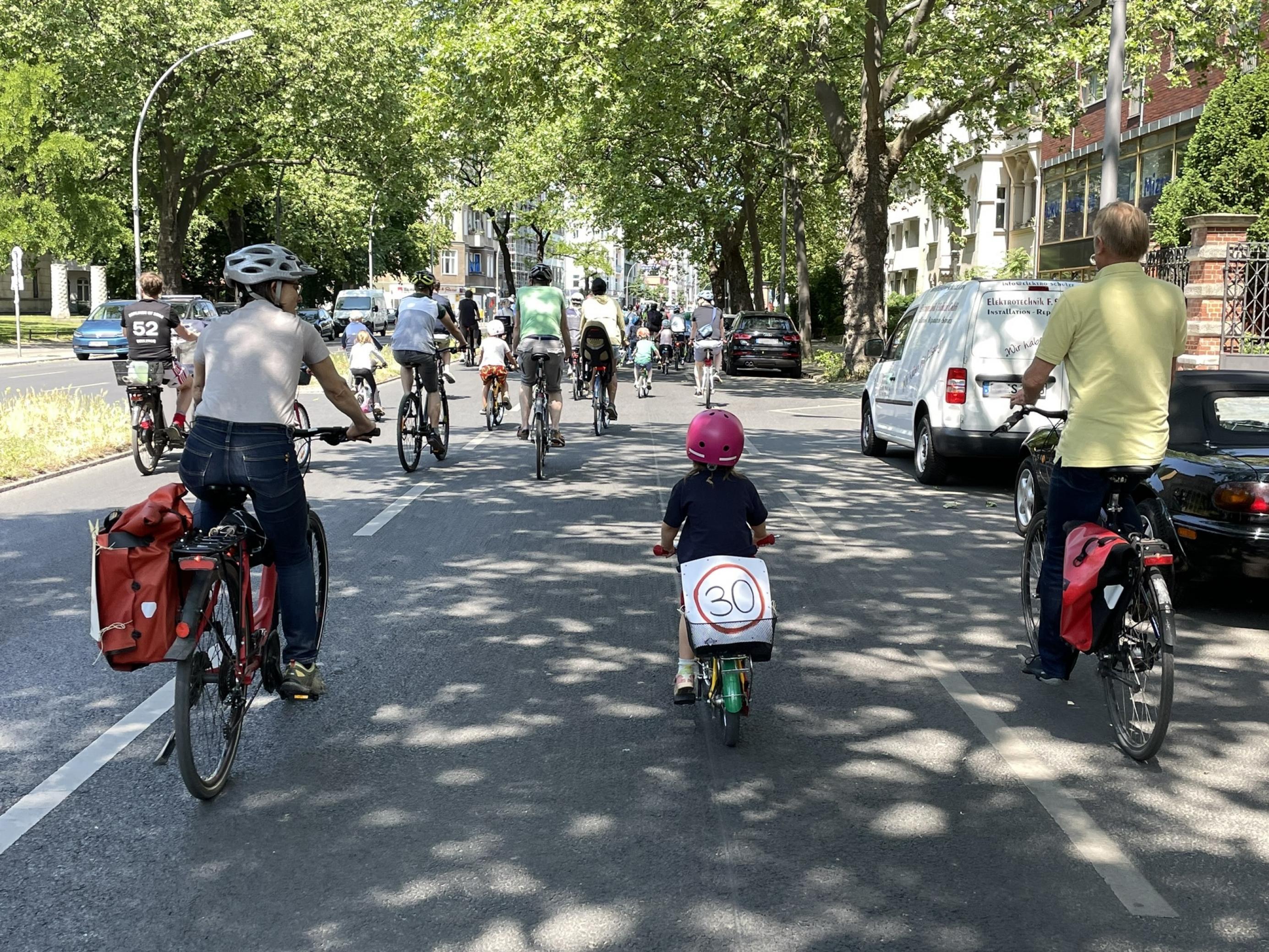 Ein Kind hat ein Schild „Tempo 30“ auf dem Gepäckträger geklemmt und radelt zwischen zwei älteren Menschen bei einer Raddemo mit.