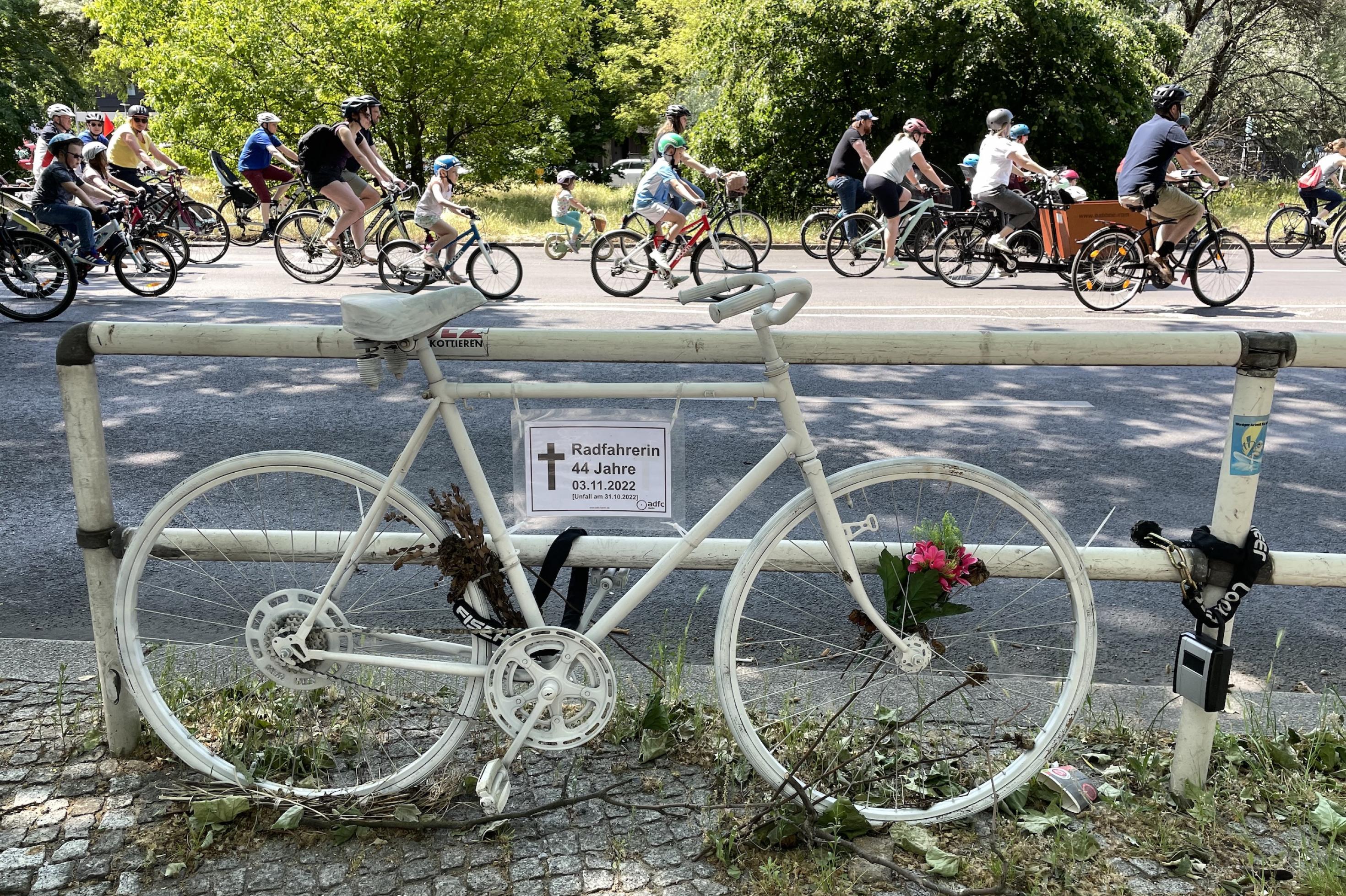 Ein weißes Fahrrad erinnert an den Unfalltod einer 44-jährigen am 3.11.2022, dahinter vorbeifahrende Radfahrer, auch Kleinkinder.