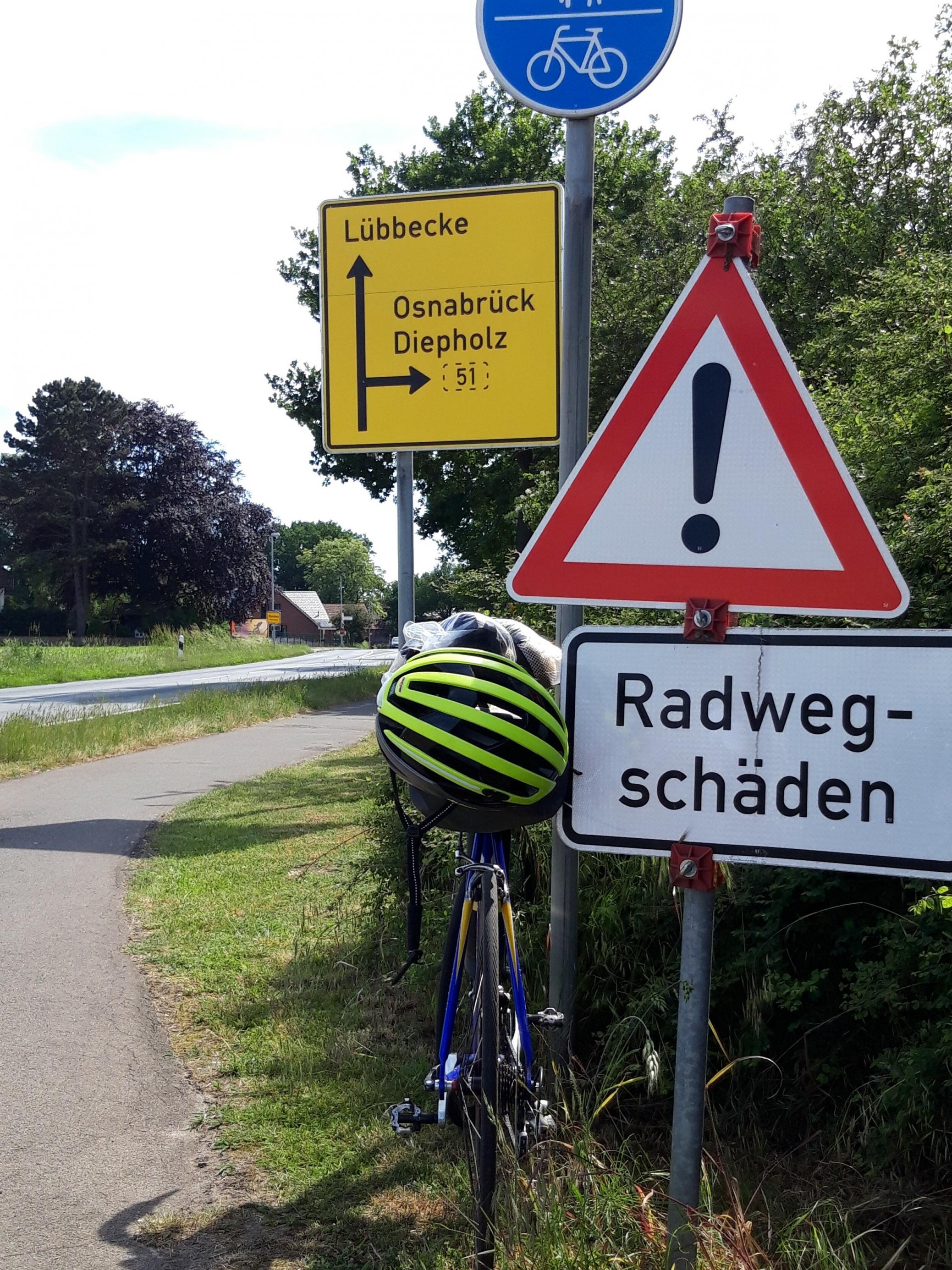 Das Vehikel des RadelndenReporters lehnt an einem Warnschild mit der Aufschrift „Radweg-Schäden“.