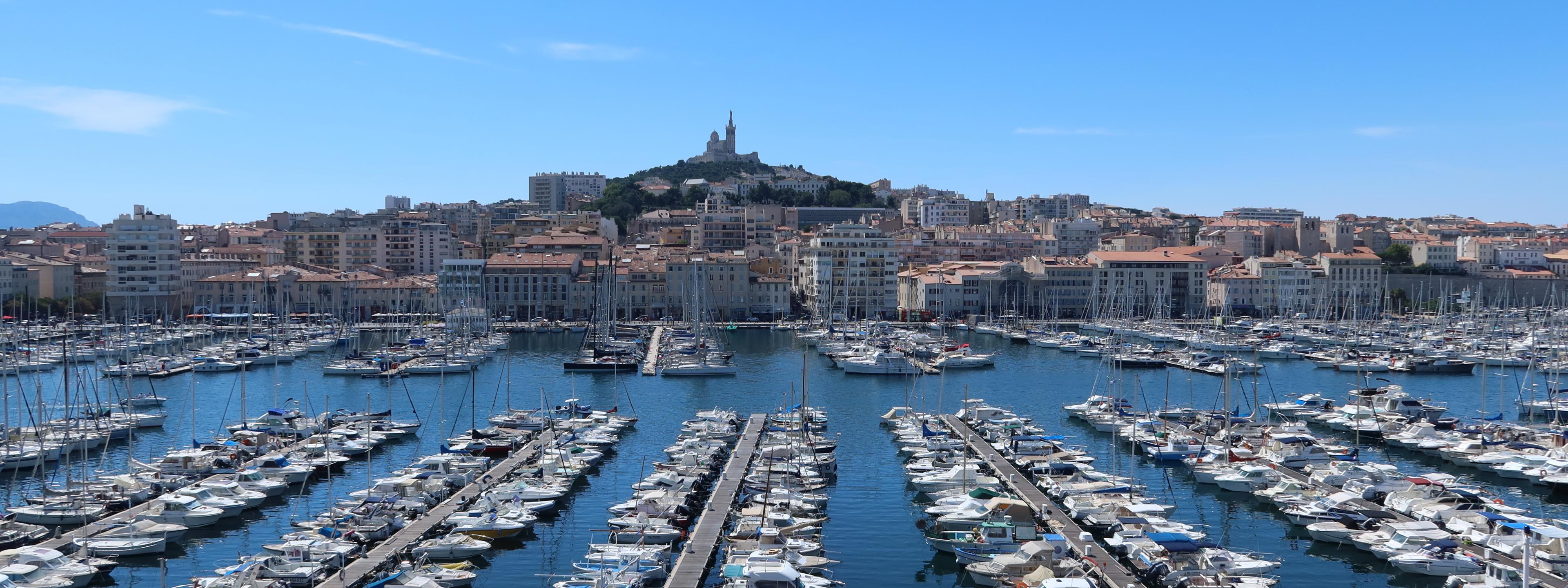 Viele Boote liegen im alten Hafen von Marseille, im Hintergrund sieht man die Kathedrale Notre-Dame de la Garde auf einem Hügel.