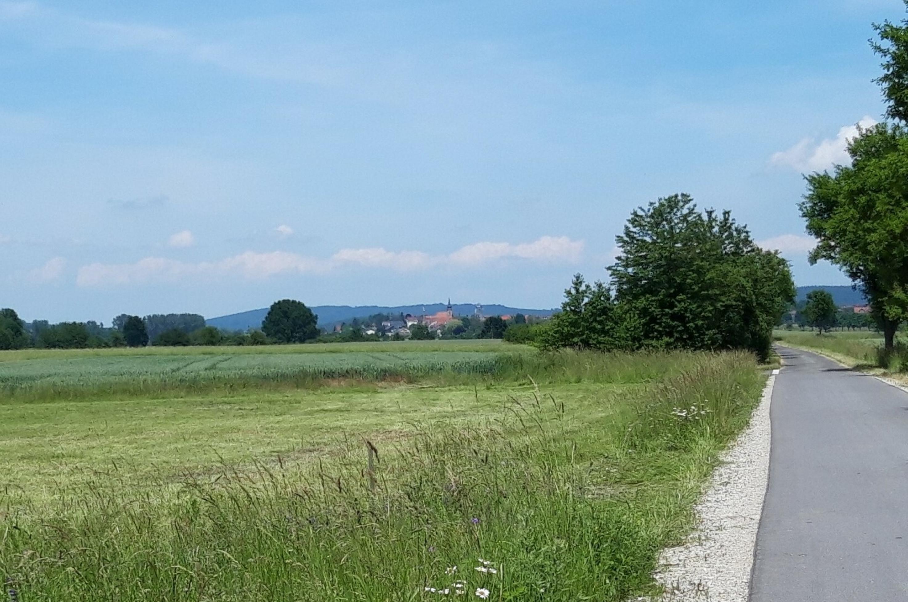 Am Horizont über Feld und Radweg heben sich vor einem Höhenzug die Dorfkirche sowie einige Häuser ab.