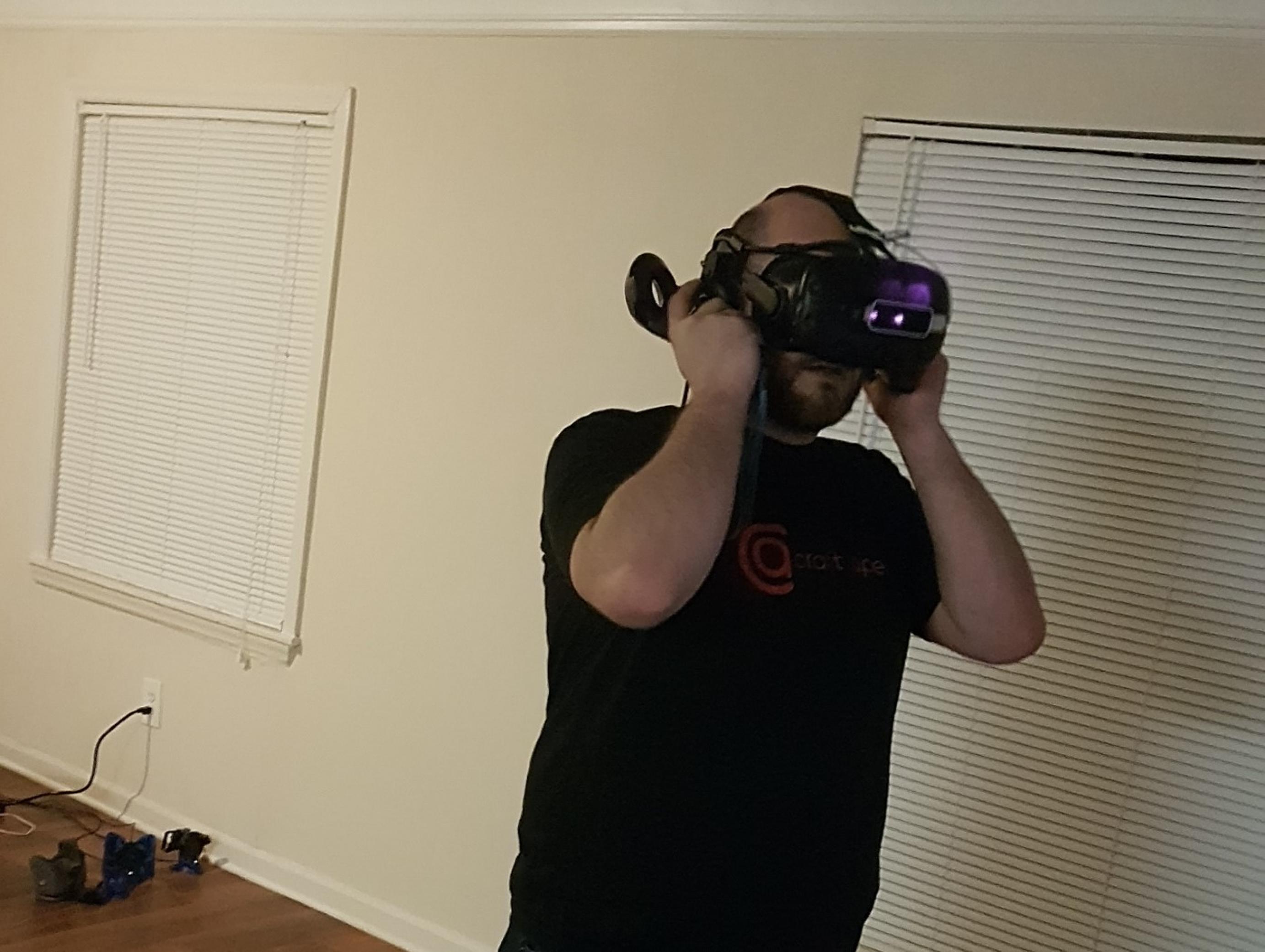Ein junger Mann mit Vollbart und beginnender Glatze trägt eineine VR-Headset auf dem Kopf. Er steht in einem leeren, kahlen Raum. Die Jalousien vor den Fenstern sind geschlossen.
