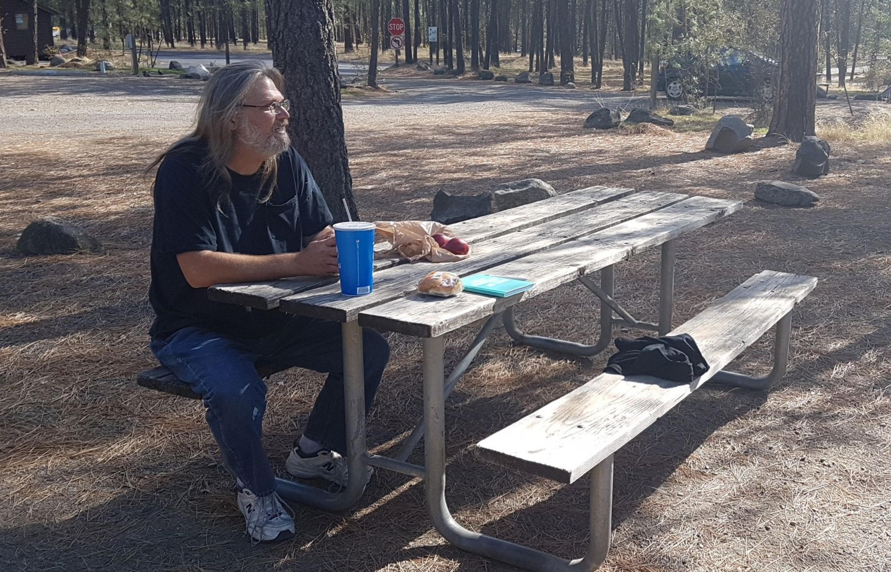 Cattz, ein älterer mann mit langen grauen Haaren, struppigem Vollbart und Brille, sitzt auf einem Picknicktisch im Wald. Die Sonne scheint ihm ins Gesicht. Vor ihm auf dem Tisch steht ein Softdrink und etwas zu Essen.