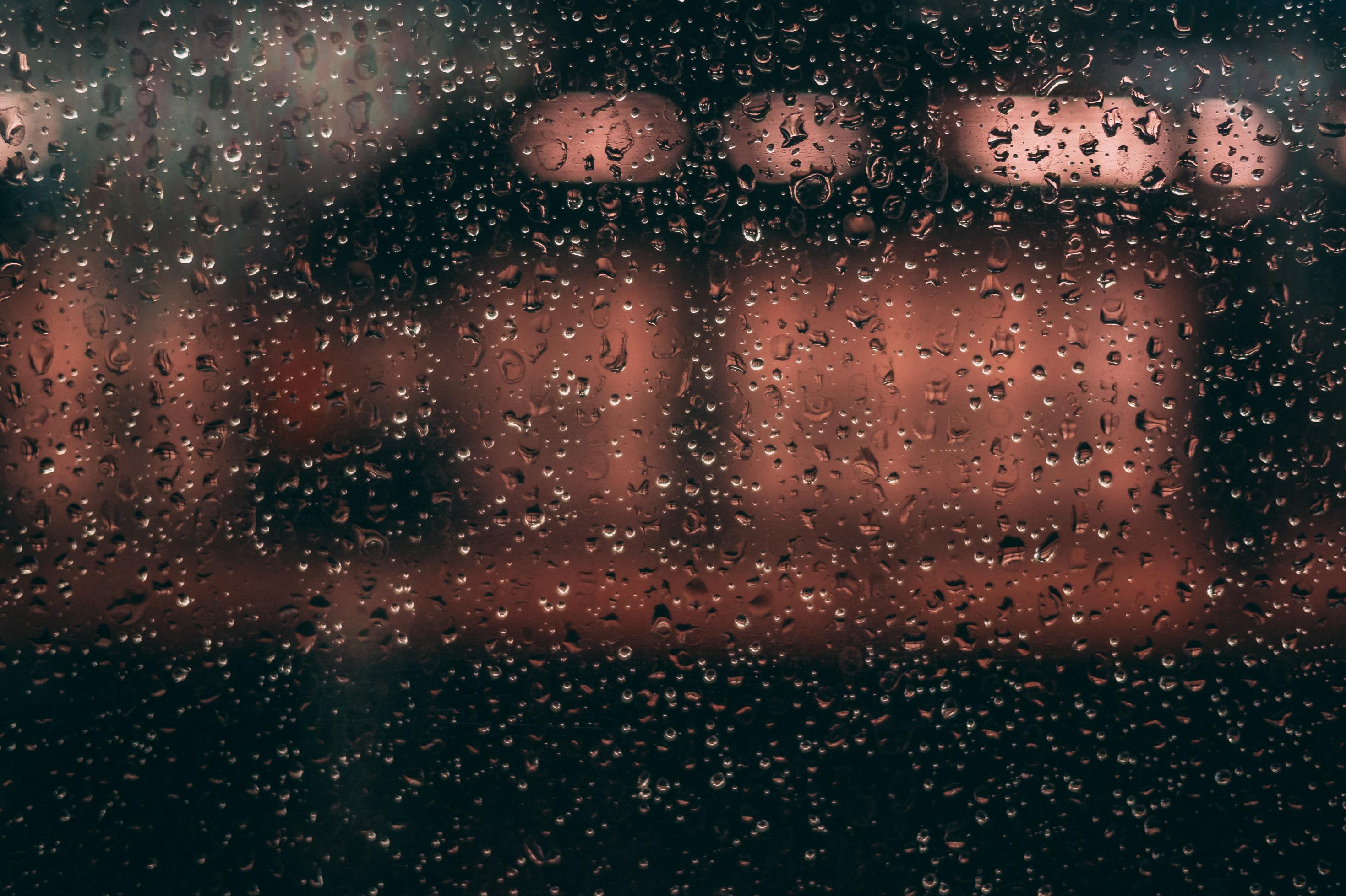 Durch eine mit Regentropfen übersäte Fensterscheibe sind unscharf die rötlichen Lichter zu erkennen, die auf einem nahen Gebäude scheinen. das Bild vermittelt das Gefühl von Isoliertheit und Abgeschiedenheit.