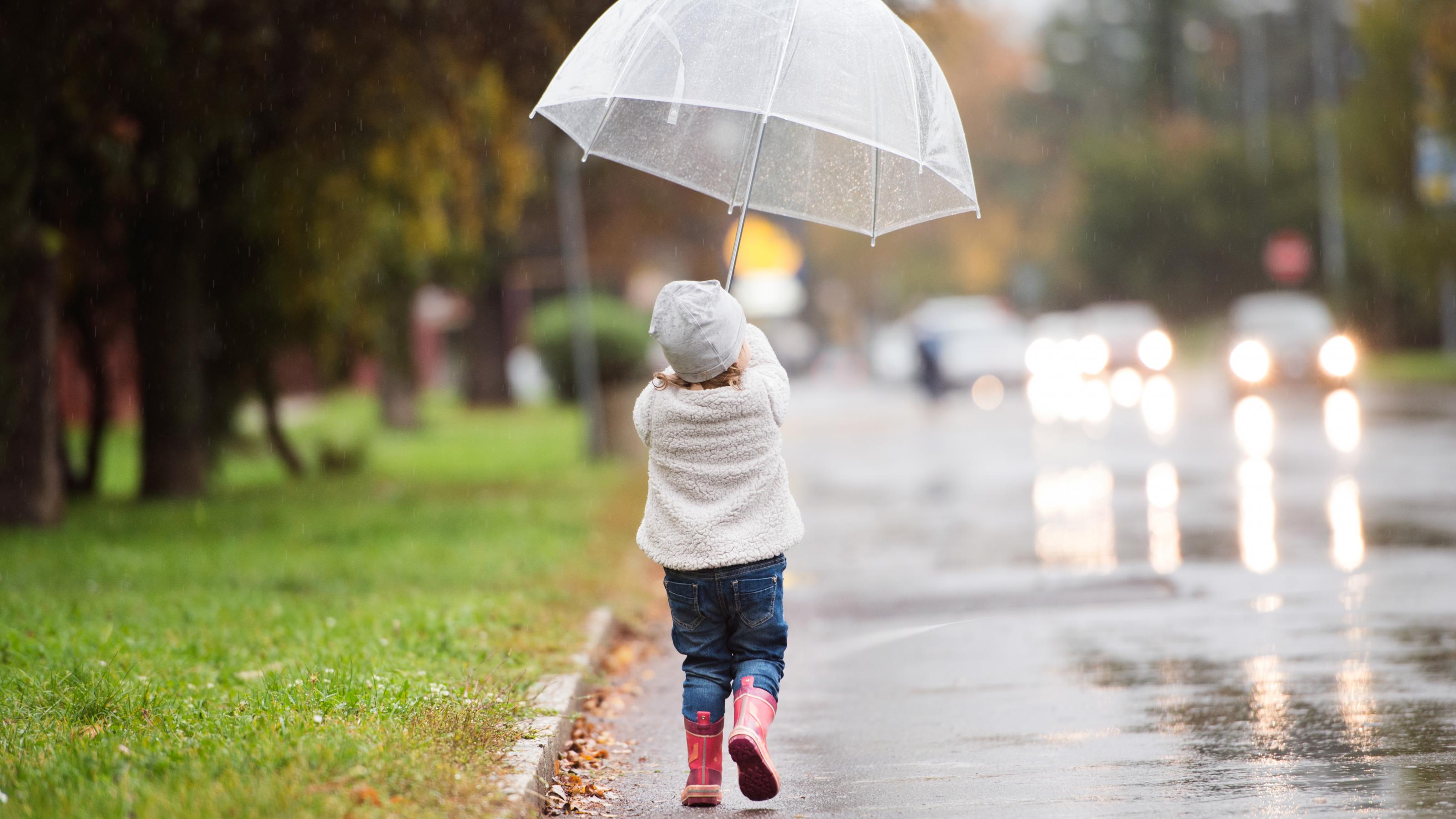Man sieht ein kleines Mädchen von hinten auf der Straße entlanggehen. Sie hält einen Schirm in der Hand, die Straße ist nass, da es gerade geregnet hat.