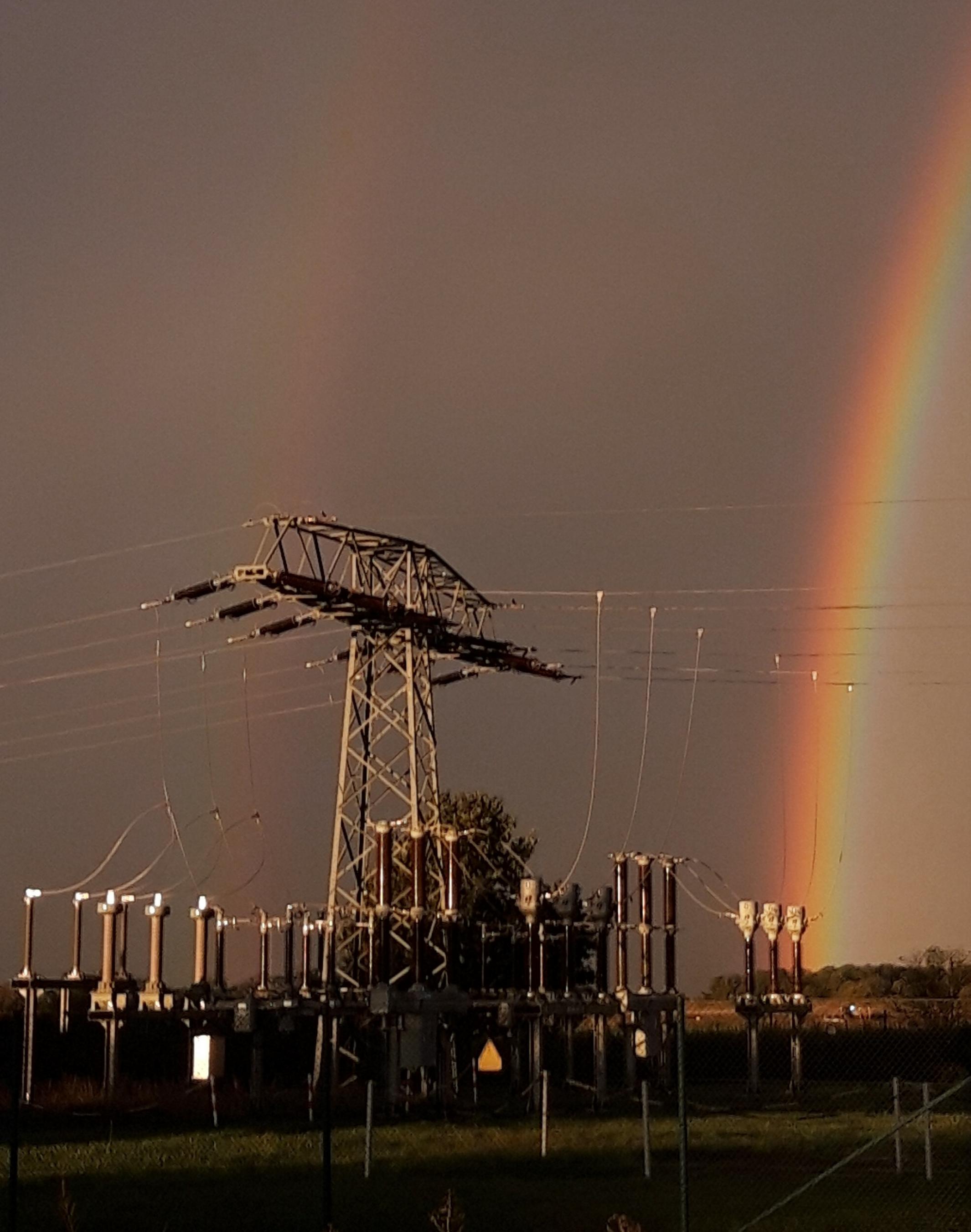 Neben den Transformatoren eines Umspannwerks strahlt vor dunklem Grau-Braun ein Regenbogen.