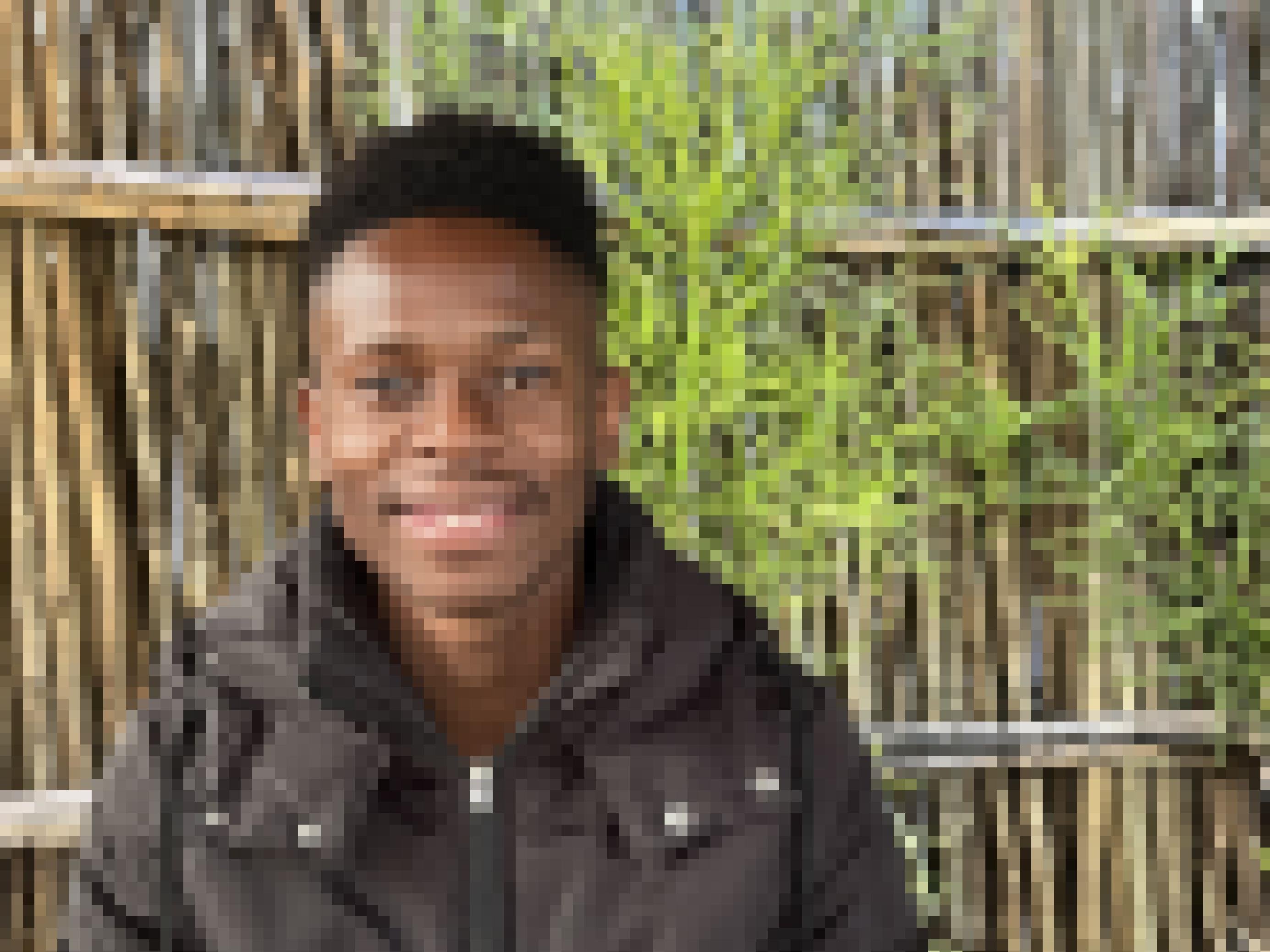 Der junge Mann lächelt in die Kamera, er sitzt in Winterjacke vor einem Bambuszaun