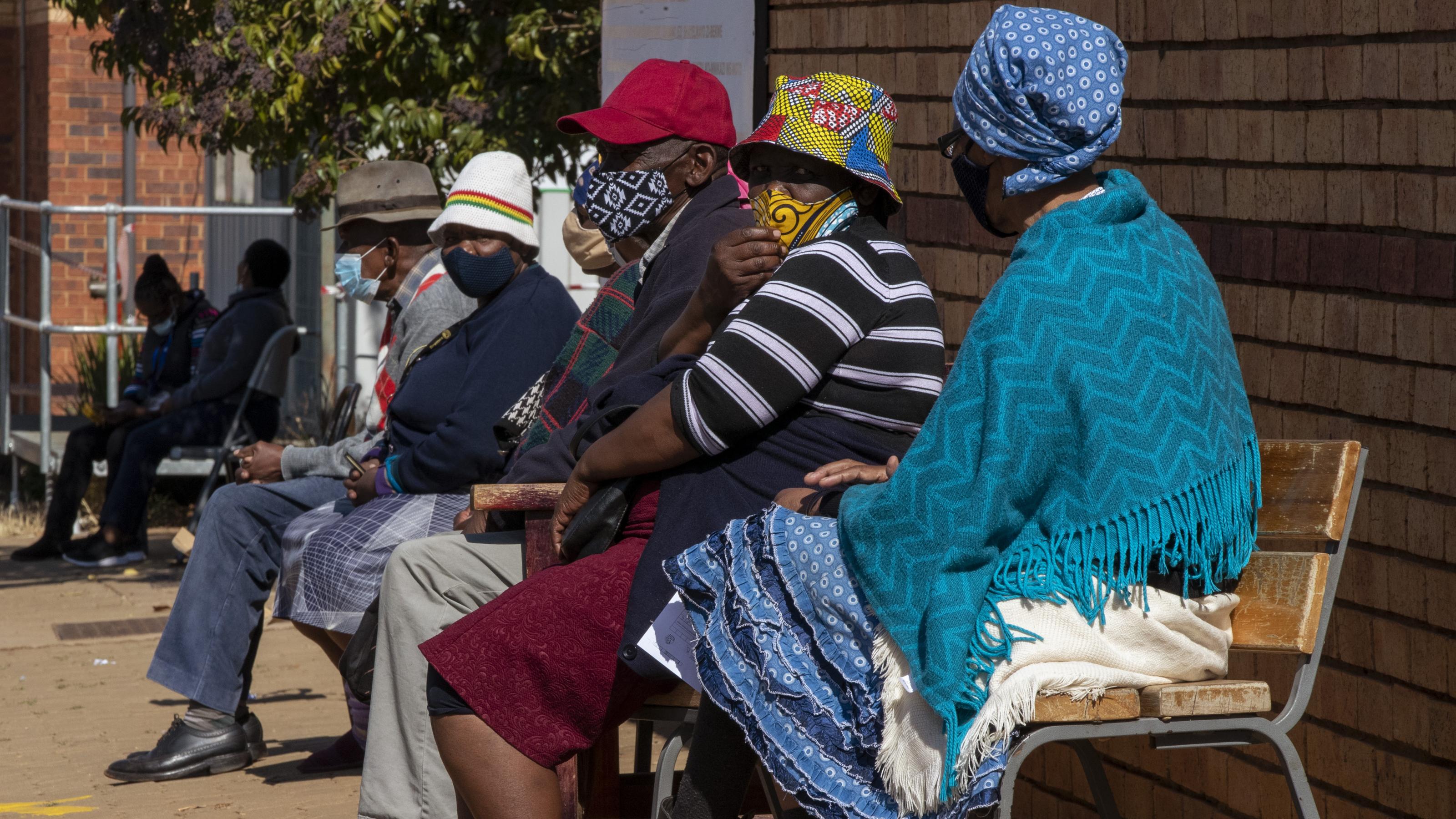 Frauen und Männer mit farbenfrohen Gesichtsmasken sitzen auf Stühlen vor der Klinik in der Schlange