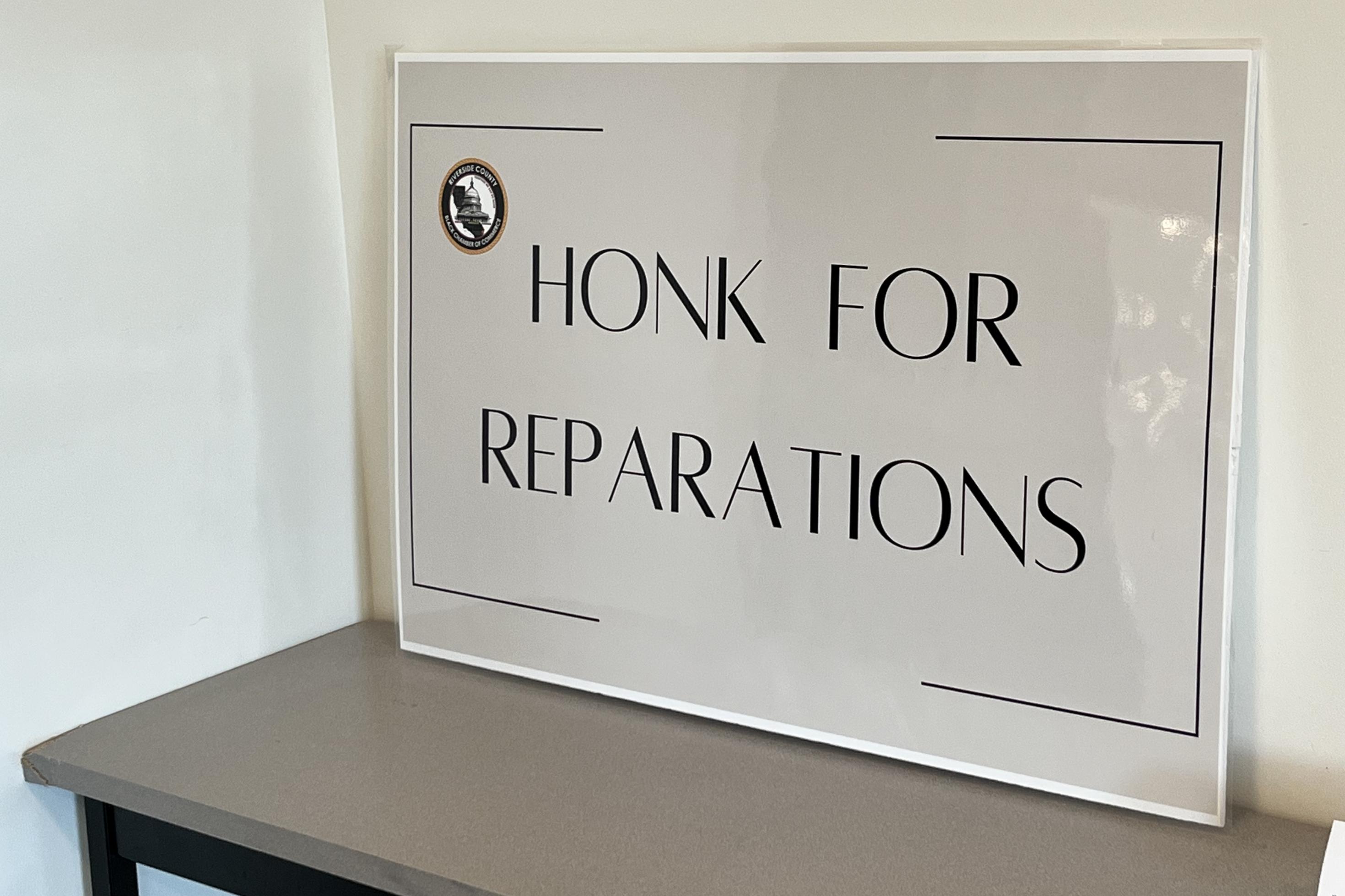 Ein weißes Schild mit schwarzer Aufschrift „Honk for reparations“ – Hupen für Entschädigung lehnt an einer weißen Wand