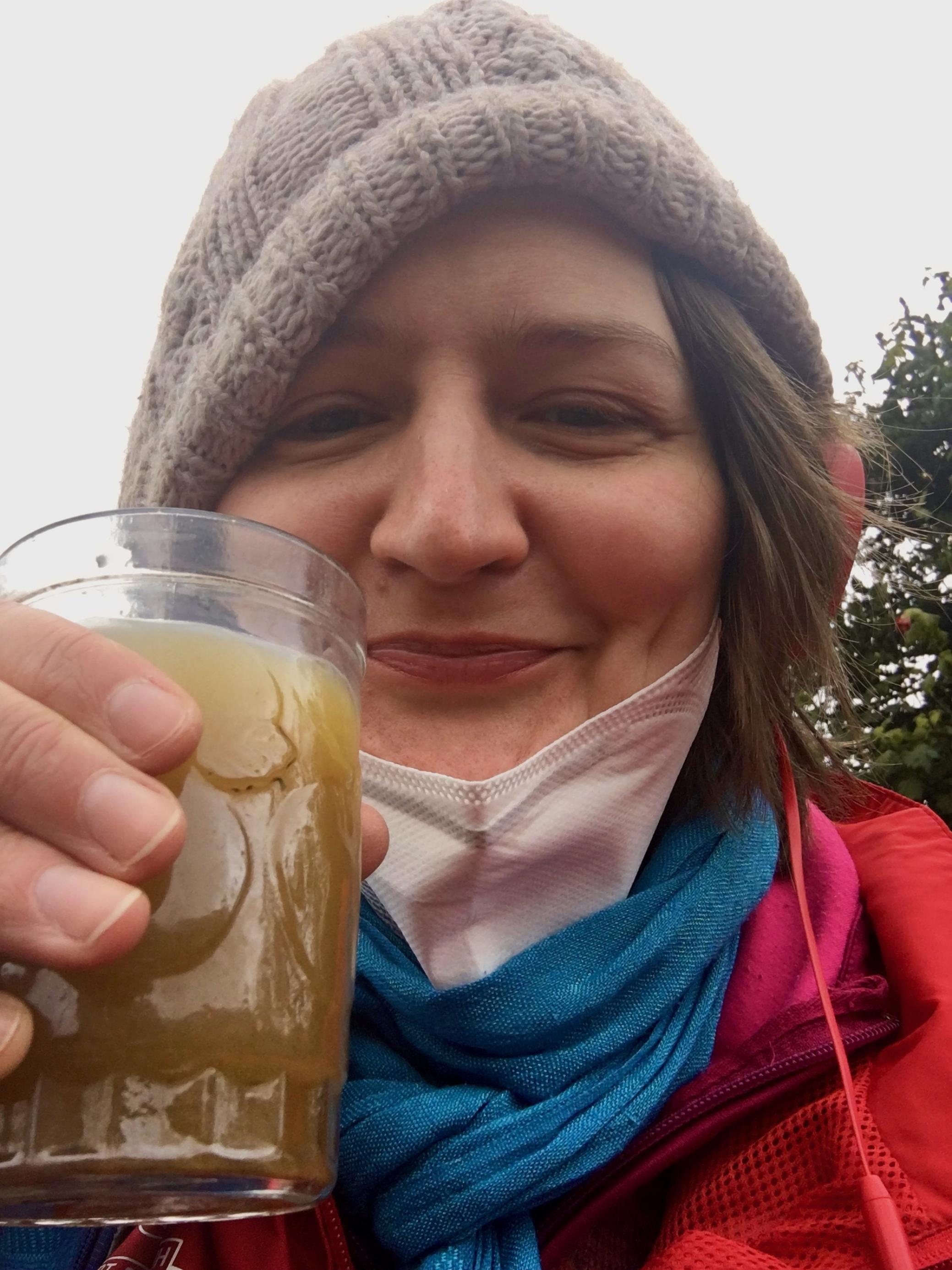 Frau mit Wollmütze, Kopfhörer und herabgezogenem Mundschutz hält Glas mit gelb-grünem Getränk mit dunklen Schwebeteilchen in die Kamera.