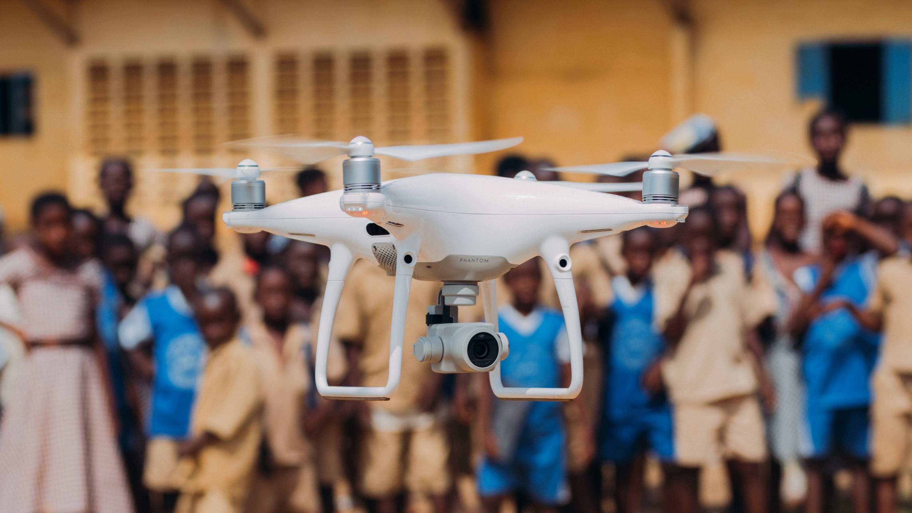 Eine Drohne schwebt im Vordergrund in der Luft. Im Hintergrund ist unscharf eine größere Gruppe afrikanischer Kinder zu sehen.