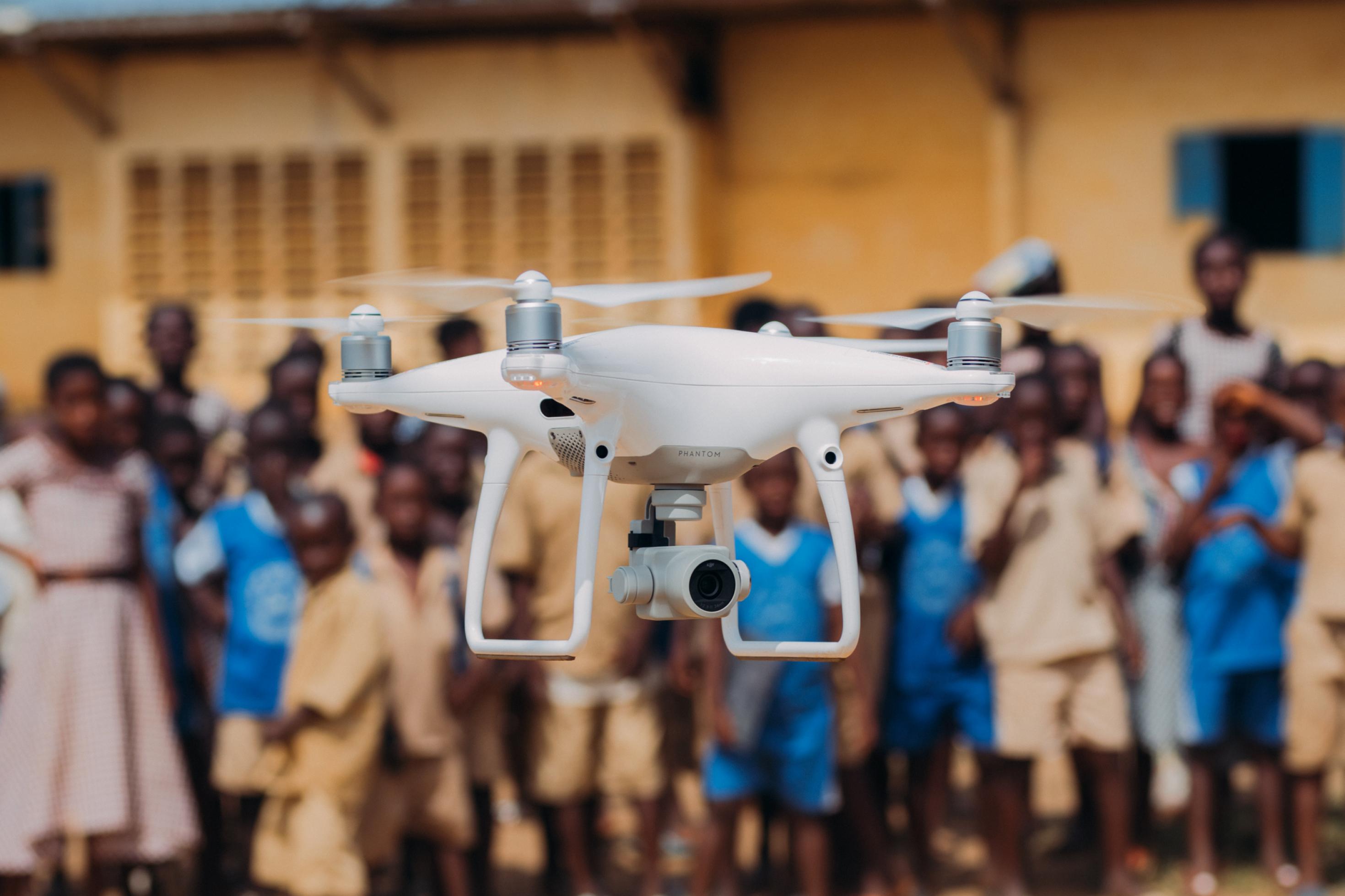 Eine Drohne schwebt im Vordergrund in der Luft. Im Hintergrund ist unscharf eine größere Gruppe afrikanischer Kinder zu sehen.