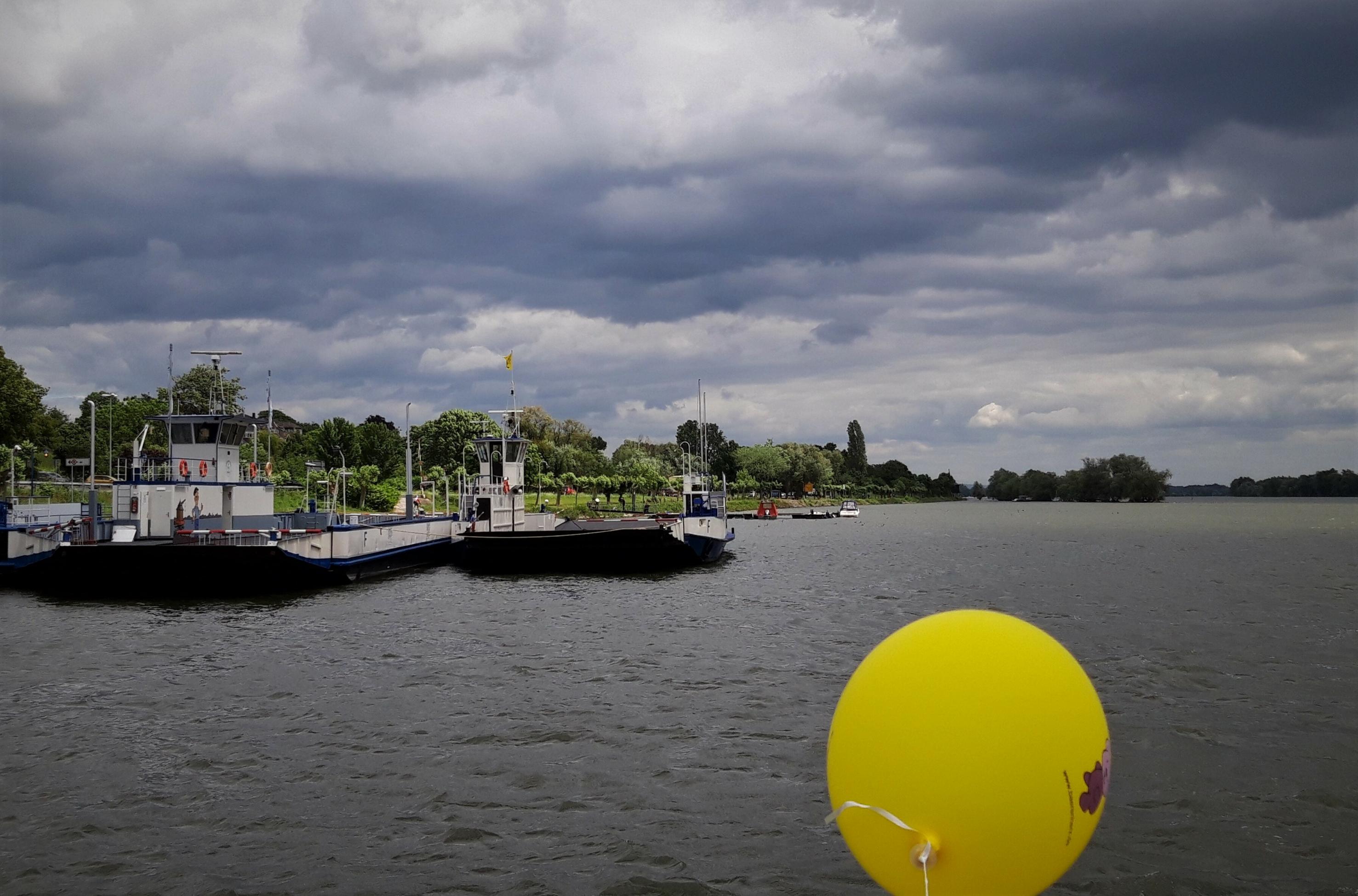 Hinter einem gelben Luftballon ist unter bleischwarzem Himmel das dunkle Rehinwasser zu sehen, auf dem eine Rheinfähre dümpelt.
