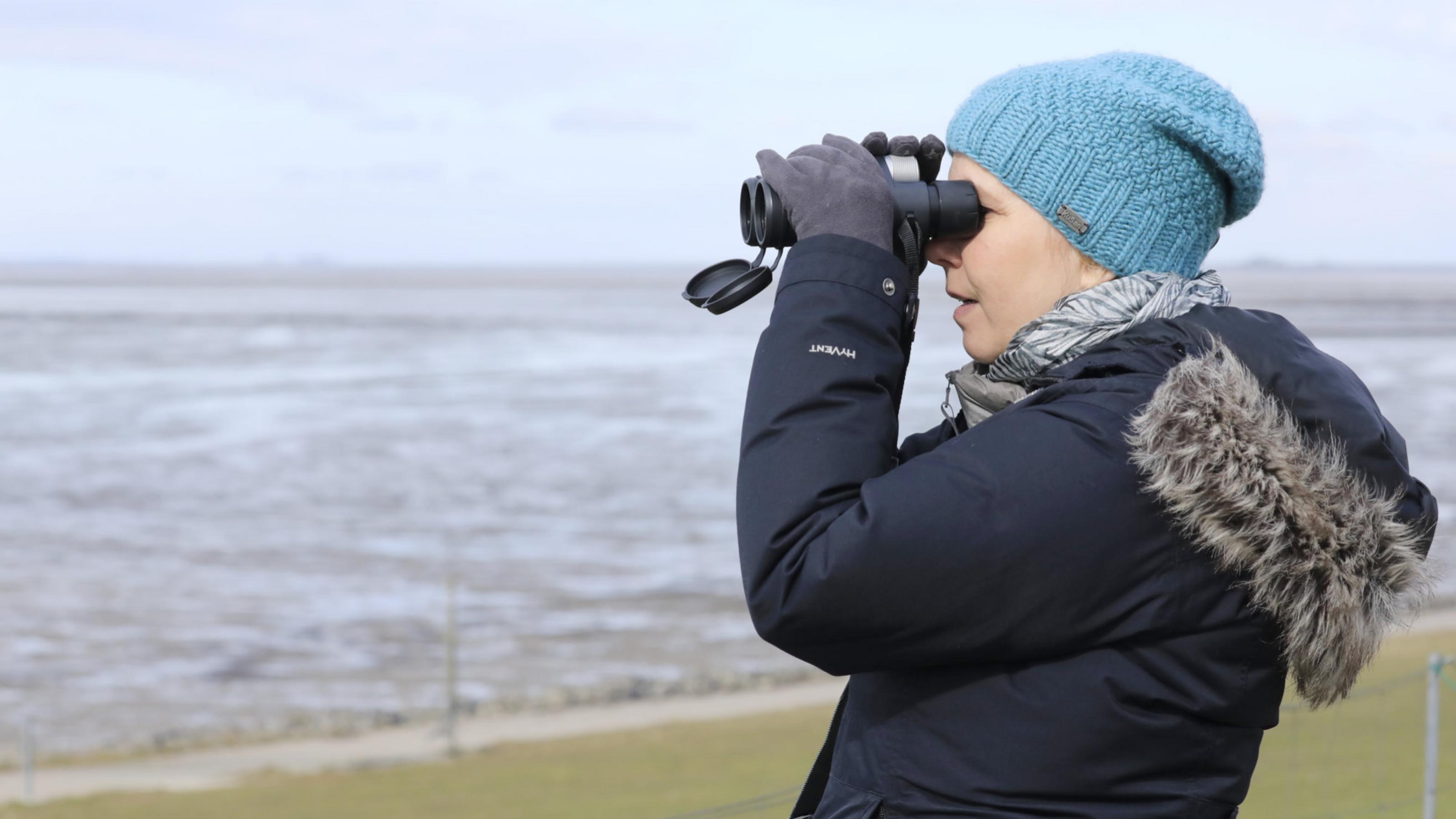 Eine Frau in Mütze und dicker Jacke schaut durch ein Fernglas. Im Hintergrund das Meer.