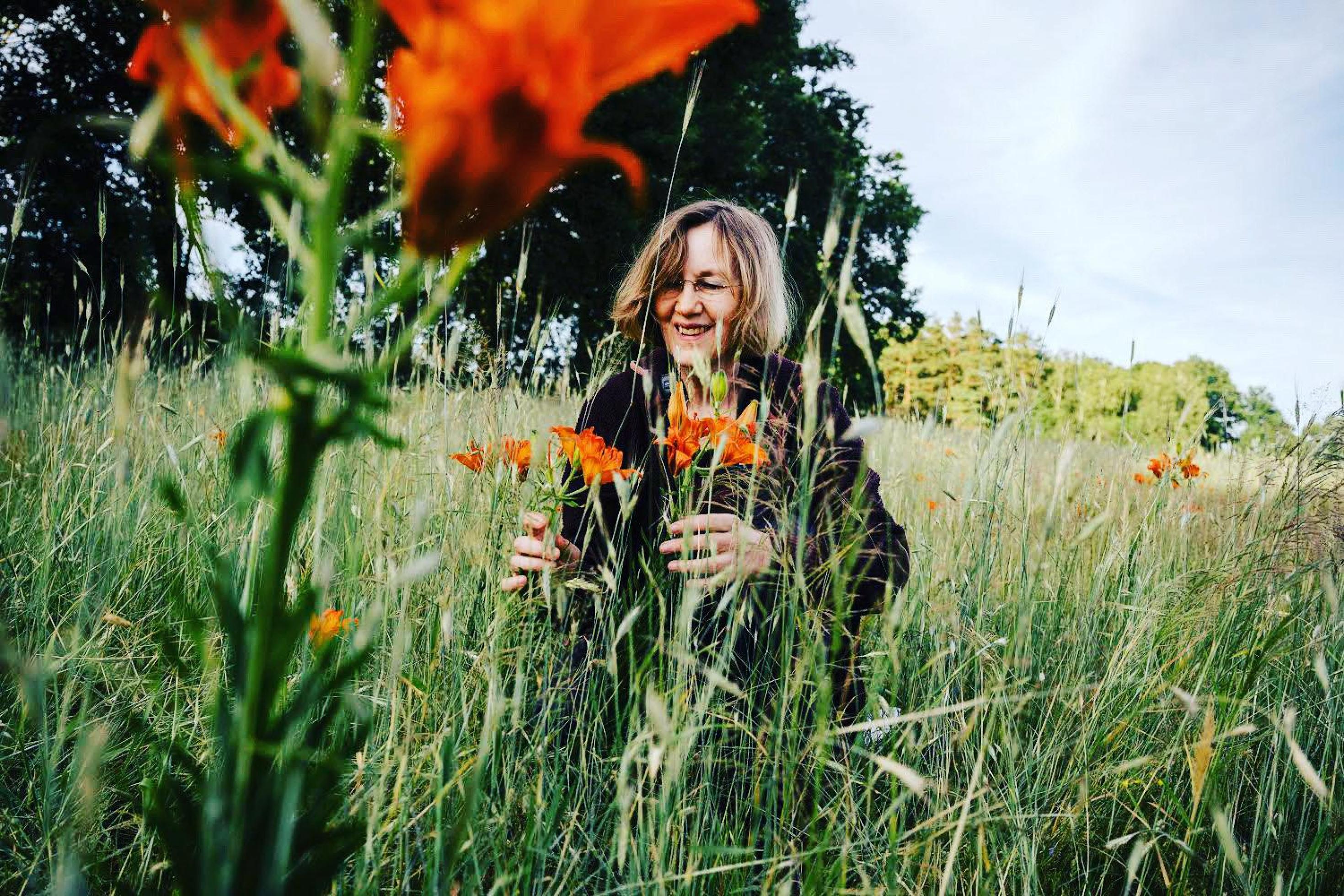 Eine Frau hockt im hohen Gras und hält eine orangefarbene Blume zwischen den Fingern. Sie lächelt.