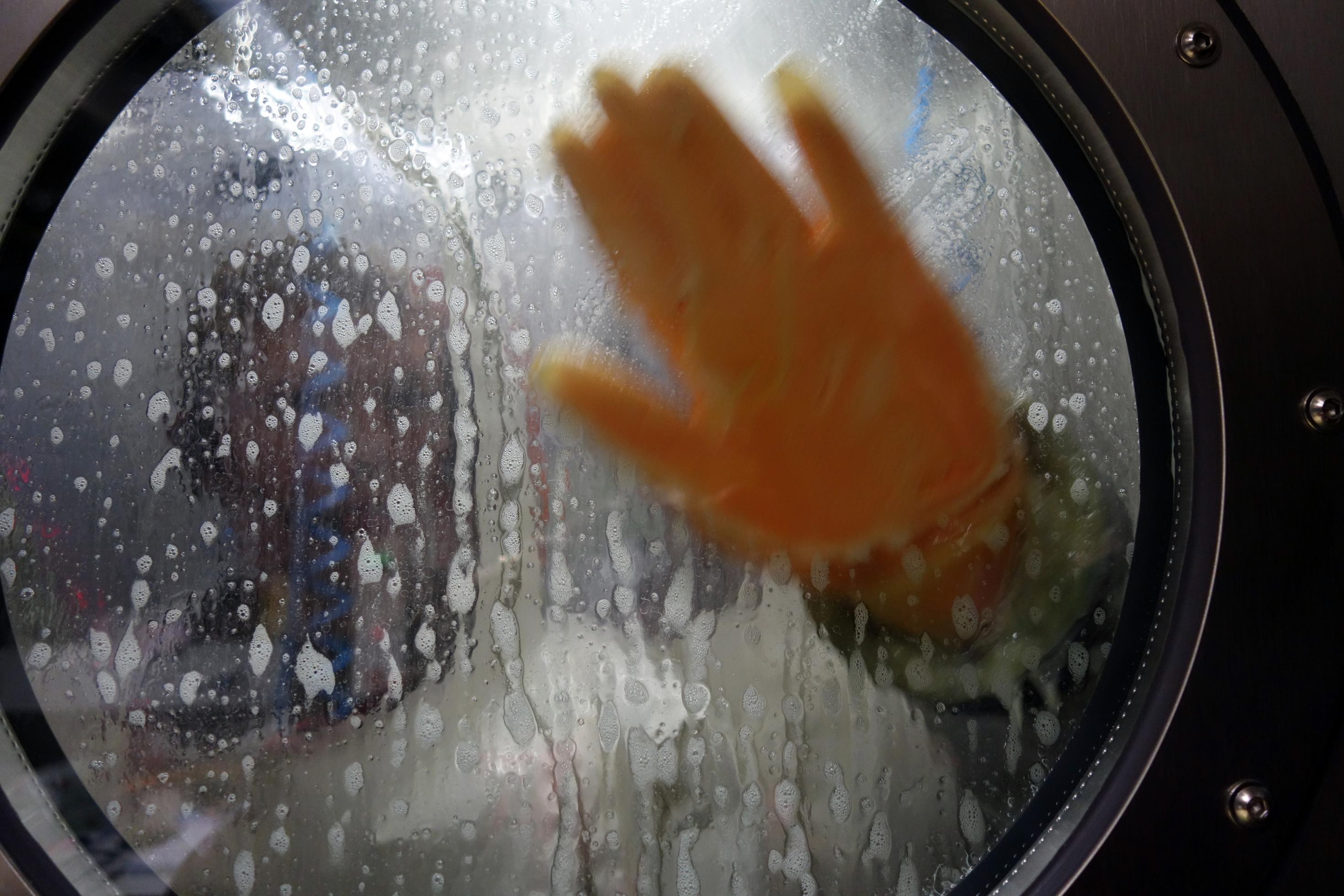 Blick durch ein rundes Fenster in die Dekontaminationsdusche des Hochsicherheitslabors am Robert Koch-Institut. Durch den Spülschaum ist zu erkennen, dass eine Person darin gerade ihren Schutzanzug abspült, eine Handfläche hält sie mit dem Schutzhandschuh an das Fenster.