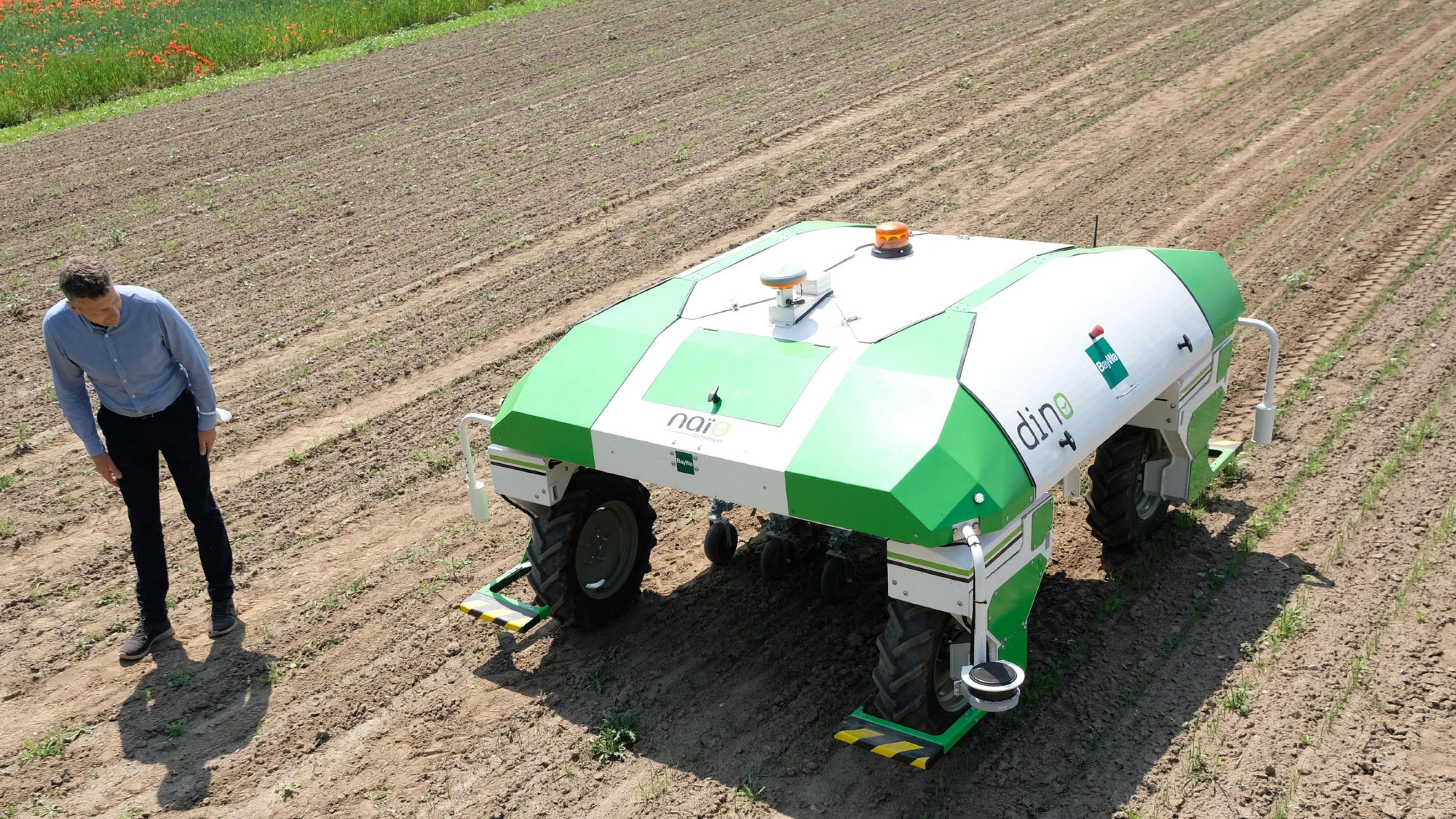 Ein grün-weißer, fahrenden Roboter unterwegs auf einem Feld. Die Maschine  bewegt sich oberhalb der Pflanzen, Zwischen den Reihen jäten automatische Greifer das Unkraut. .