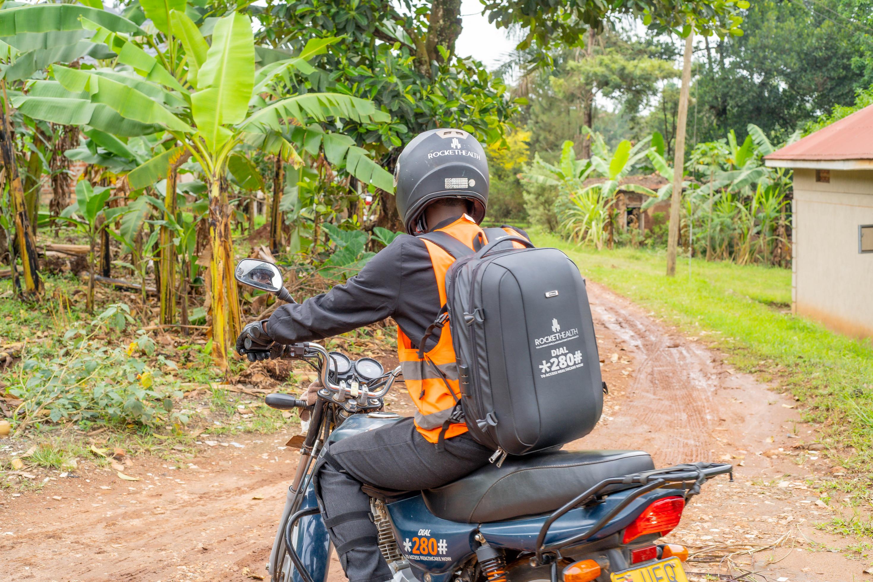 Der Motorradfahrer ist von hinten zu sehen, er trägt einen Rocket Health Rucksack, vor ihm eine ungeteerte Straße enlang Bananenfeldern
