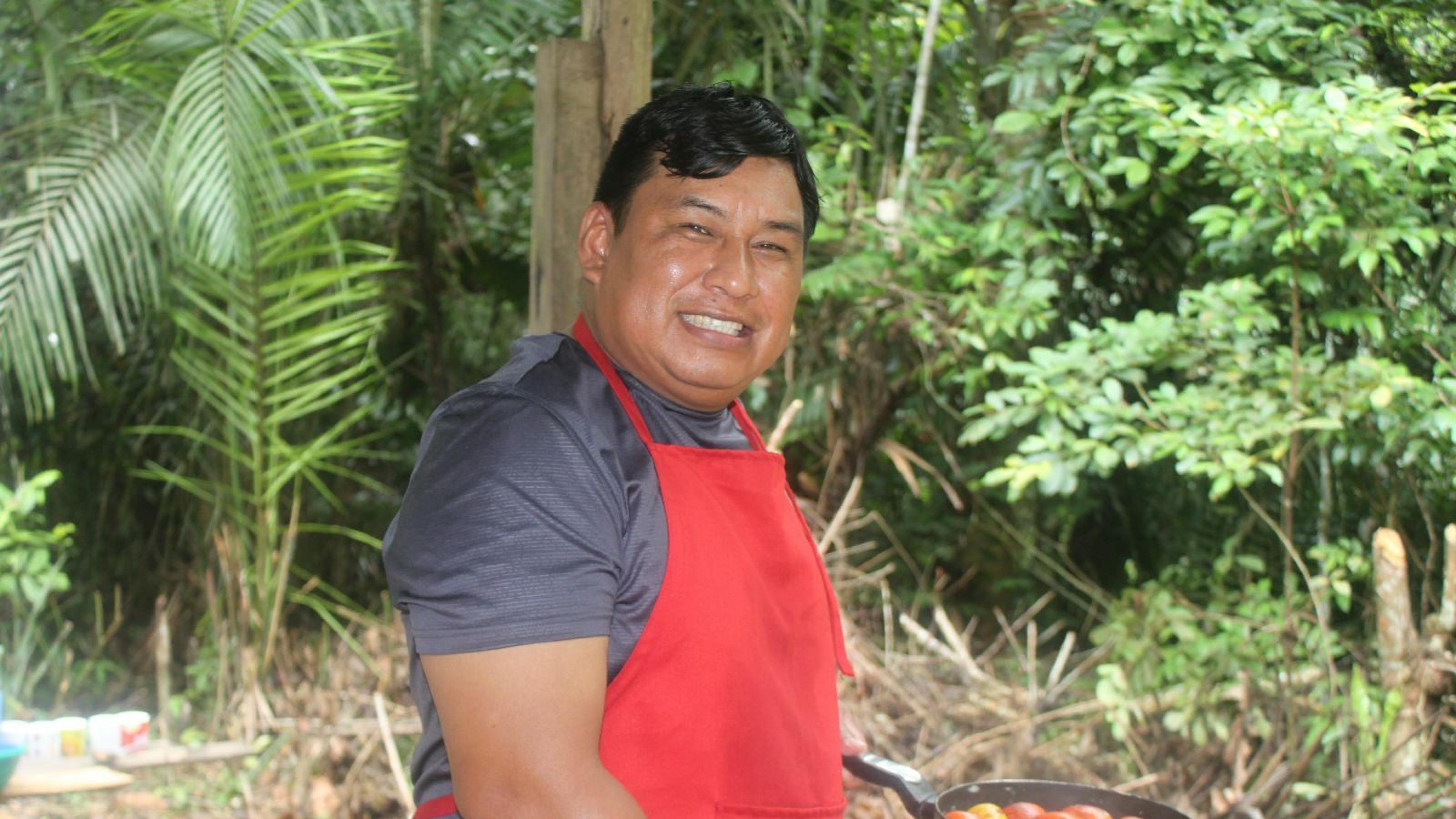 Mann, Ende dreißig, dunkle Haare, braunes Gesicht, trägt eine rote Schürze und hält eine Pfanne mit roten Früchten in der Hand. Im Hintergrund Regenwald.