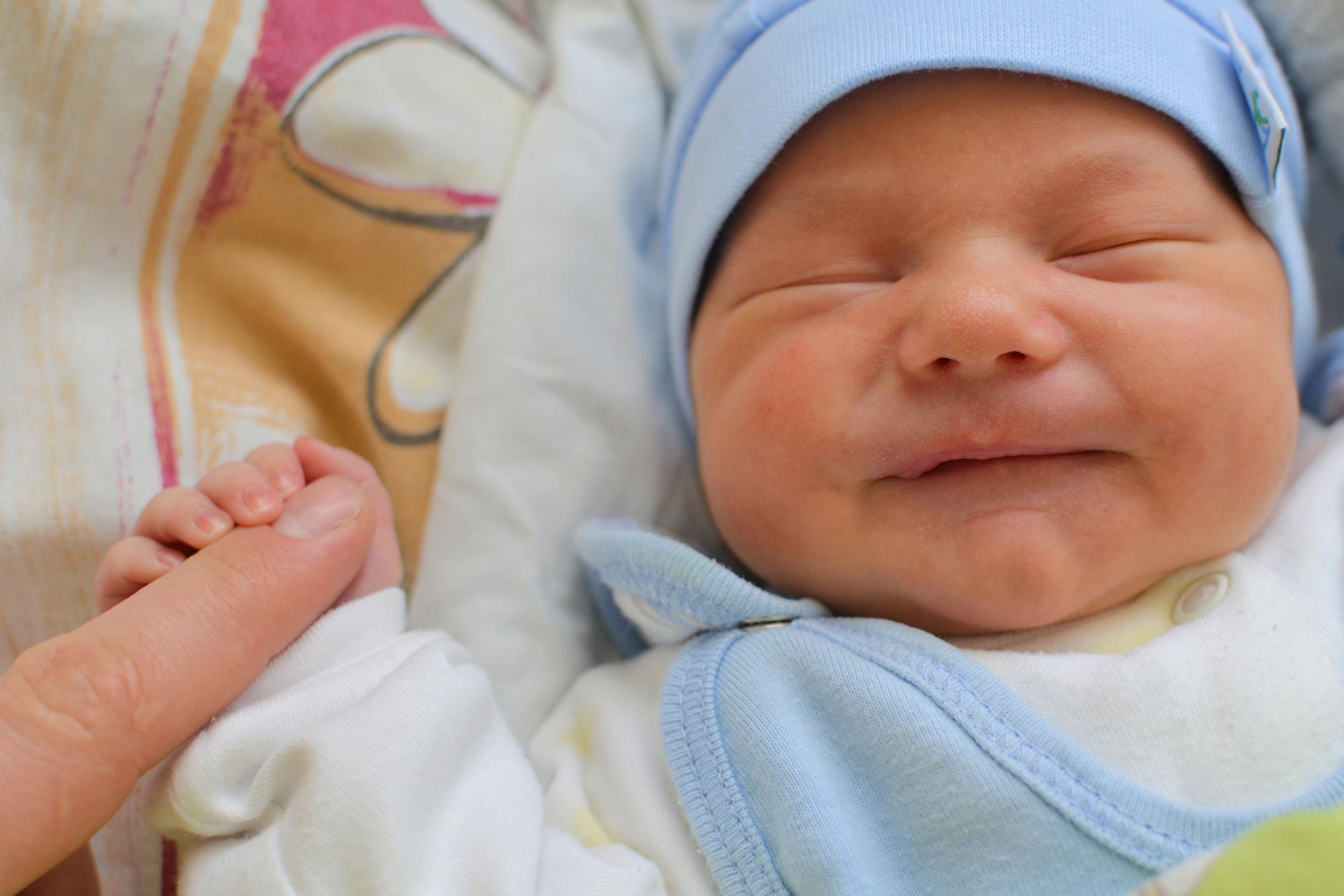 Ein neugeborenes Baby mit geschlossenen Augen und einer blauen Mütze umfasst den Zeigefinger eines Erwachsenen.