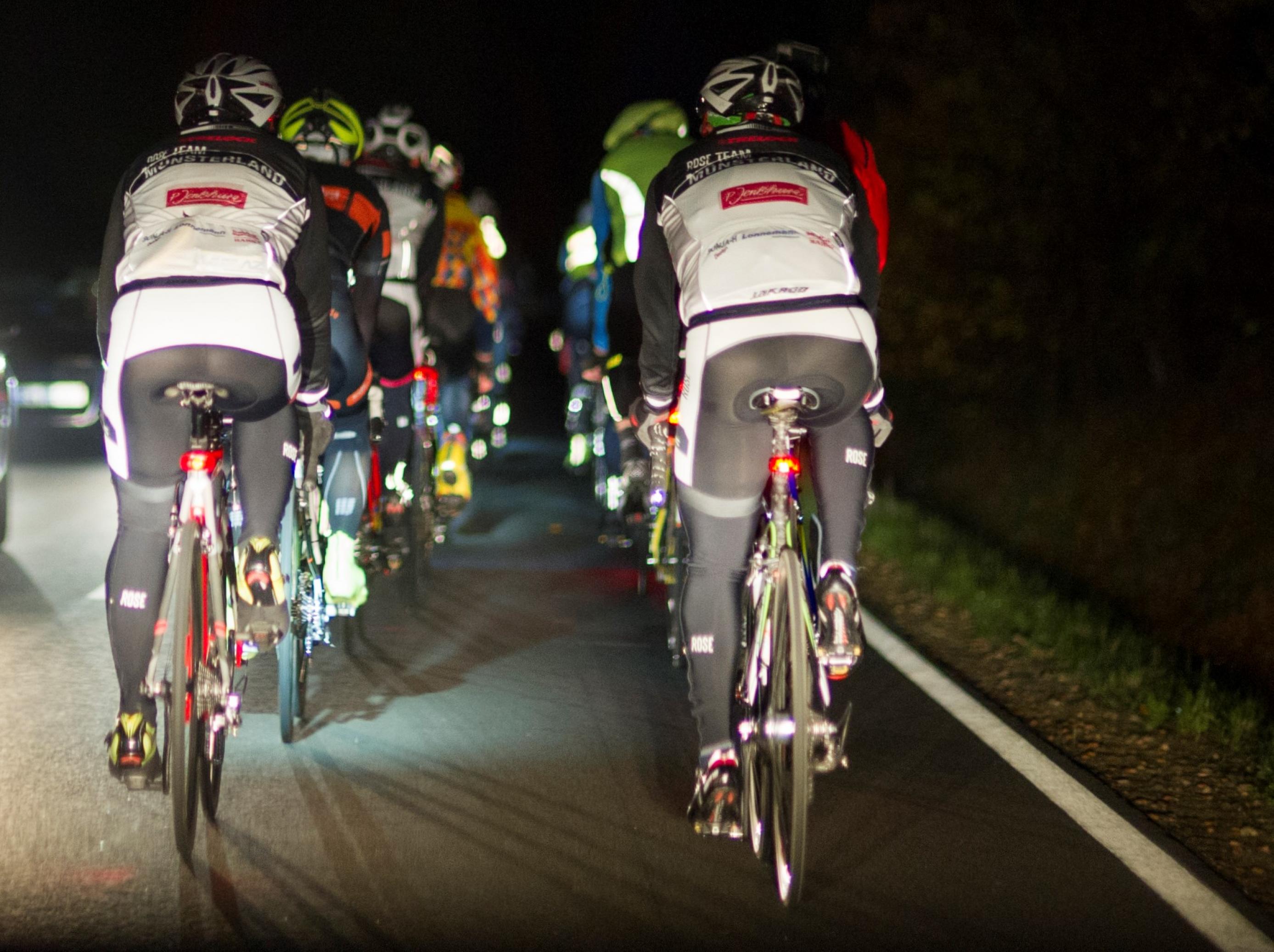 Eine Gruppe Rennradfahrer, im Dunkeln von hinten auf einer Landstraße fotografiert.
