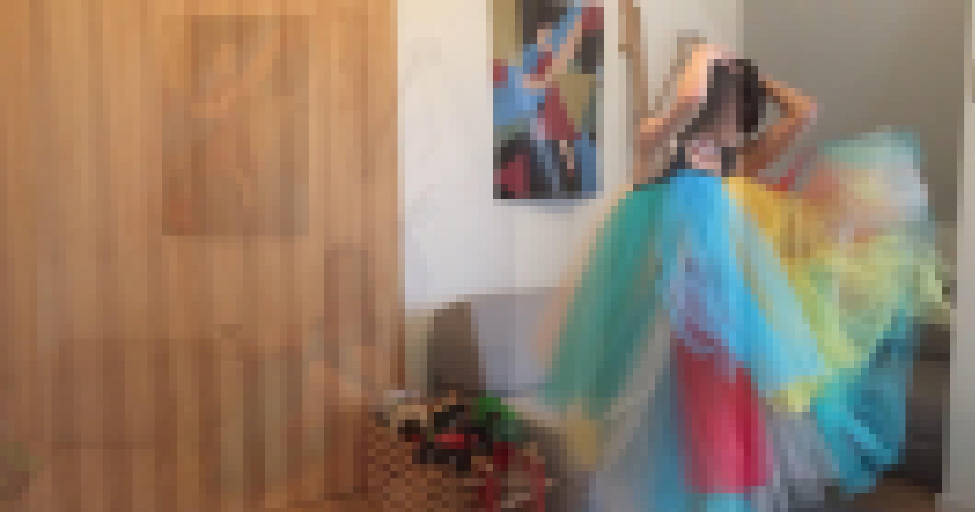 Filmstill aus dem „Gaia-Projekt“ des Theaterduos kainkollektiv: Die in Berlin und Kanada lebende Tänzerin Catherine Jodoin steht in ihrem Wohnzimmer in einem bunten Tutu und steckt sich die Haare hoch. Hinter ihr ein Poster mit einer Tänzerin. Die linke Bildhälfte nimmt eine Schrankwand ein. Daneben steht hinter ihr ein Sofa, an der Seite eine Kiste mit Spielzeug.