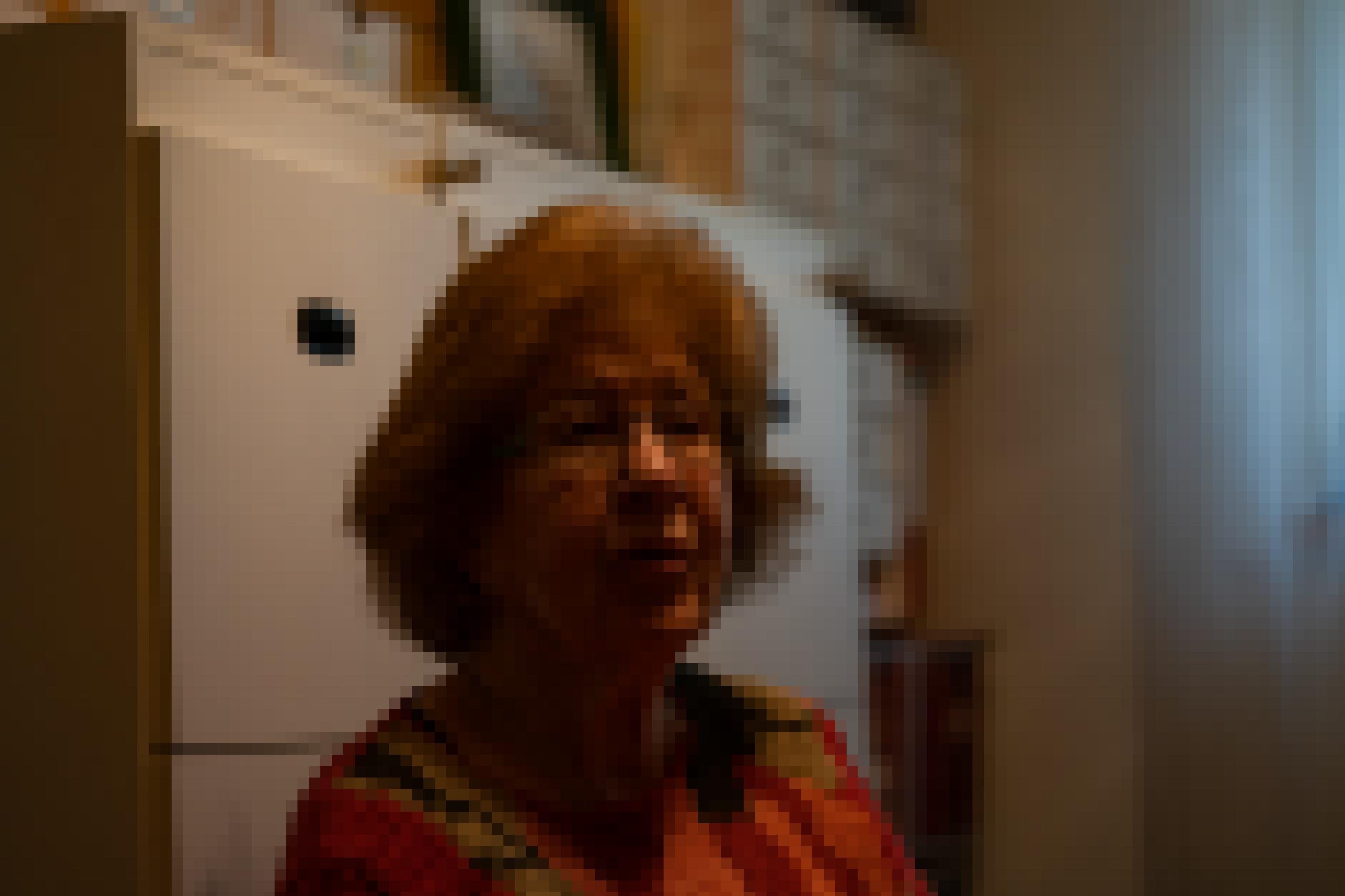 Das Gesicht vonRuth Gross trägt einen bunten Pullover und Brille. Im Hintergrund ist eine Wand mit Regalen und Boxen zu sehen, in denen Bilder ihres Vaters liegen.