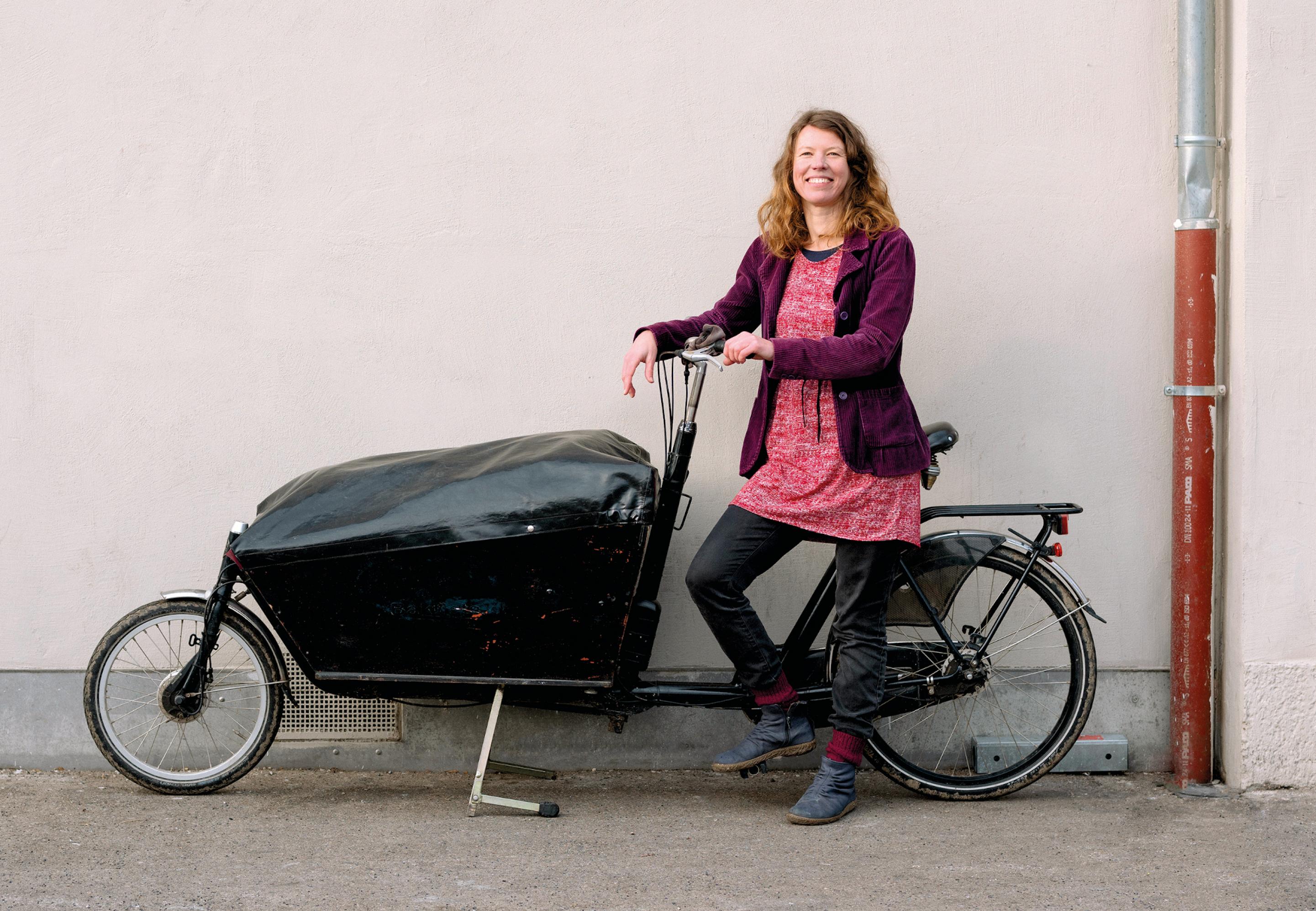 Eine Frau mit pinkem Kleid, dunkelblauer Jeans und lila Jacke steht lachend an einem Lastenrad. Dessen Ladefläche ist voll und mit einer schwarzen Plane abgedeckt.