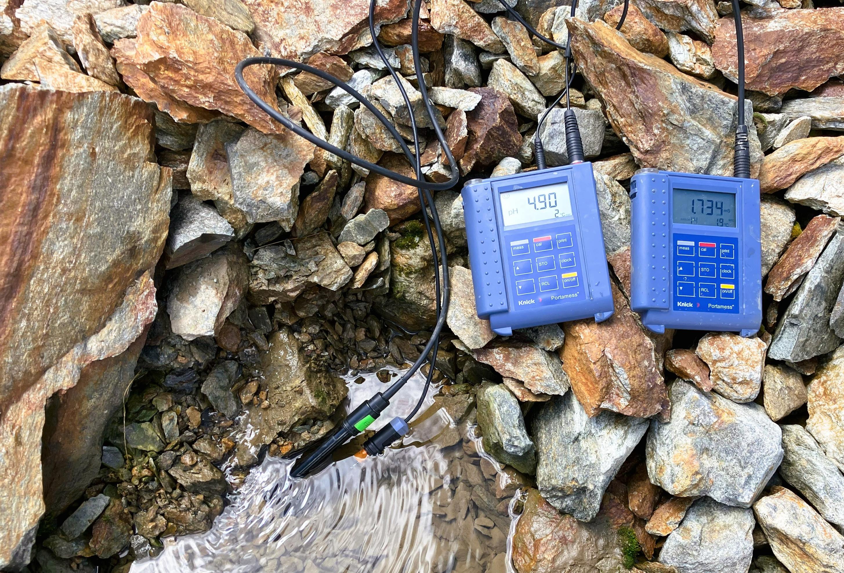 Zwei handliche blaue Elektrogeräte, von denen Kabel in eine Wasserlache führen, liegen auf losem braunem Gestein.