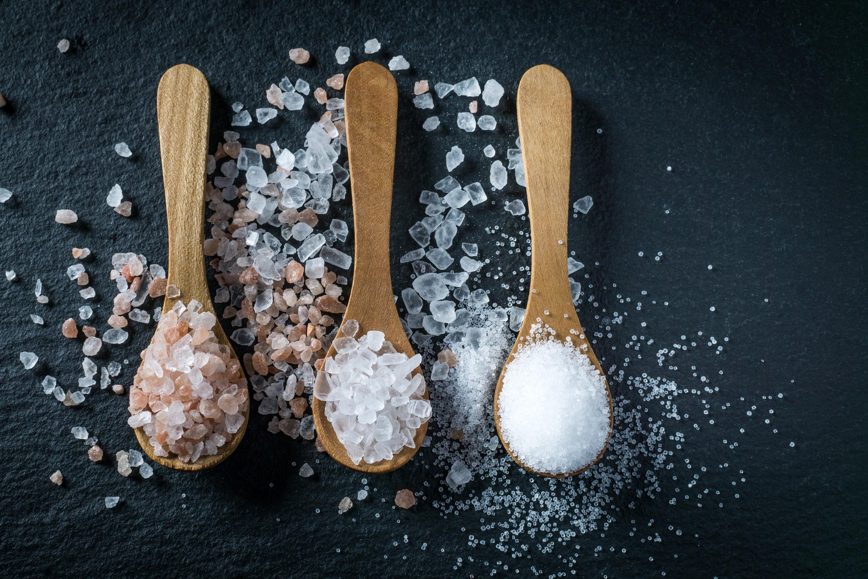 Draufsicht: Schwarzer Hintergrund, darauf liegen drei Holzlöffel gefüllt mir grobkörnigem Salz: Himalaya-Salz, Meersalz und fein gekörntes jodiertes Salz.