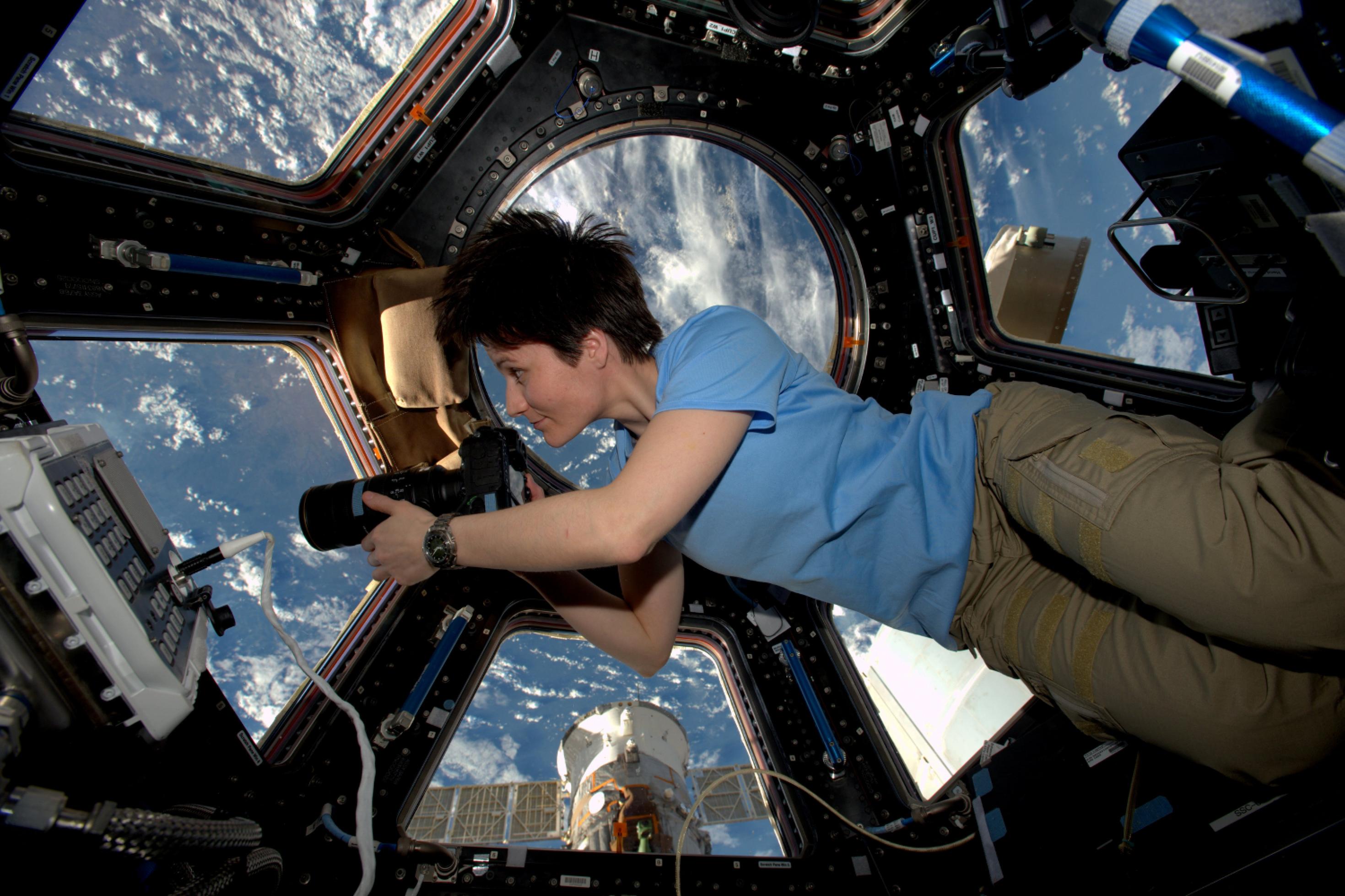 ESA-Astronautin Samantha Cristoforetti schwebt im Cupula-Modul der ISS, mit großen Fenstern und Erde im Hintergrund, hält eine große Kamera in der Hand und blickt konzentriert nach draußen.