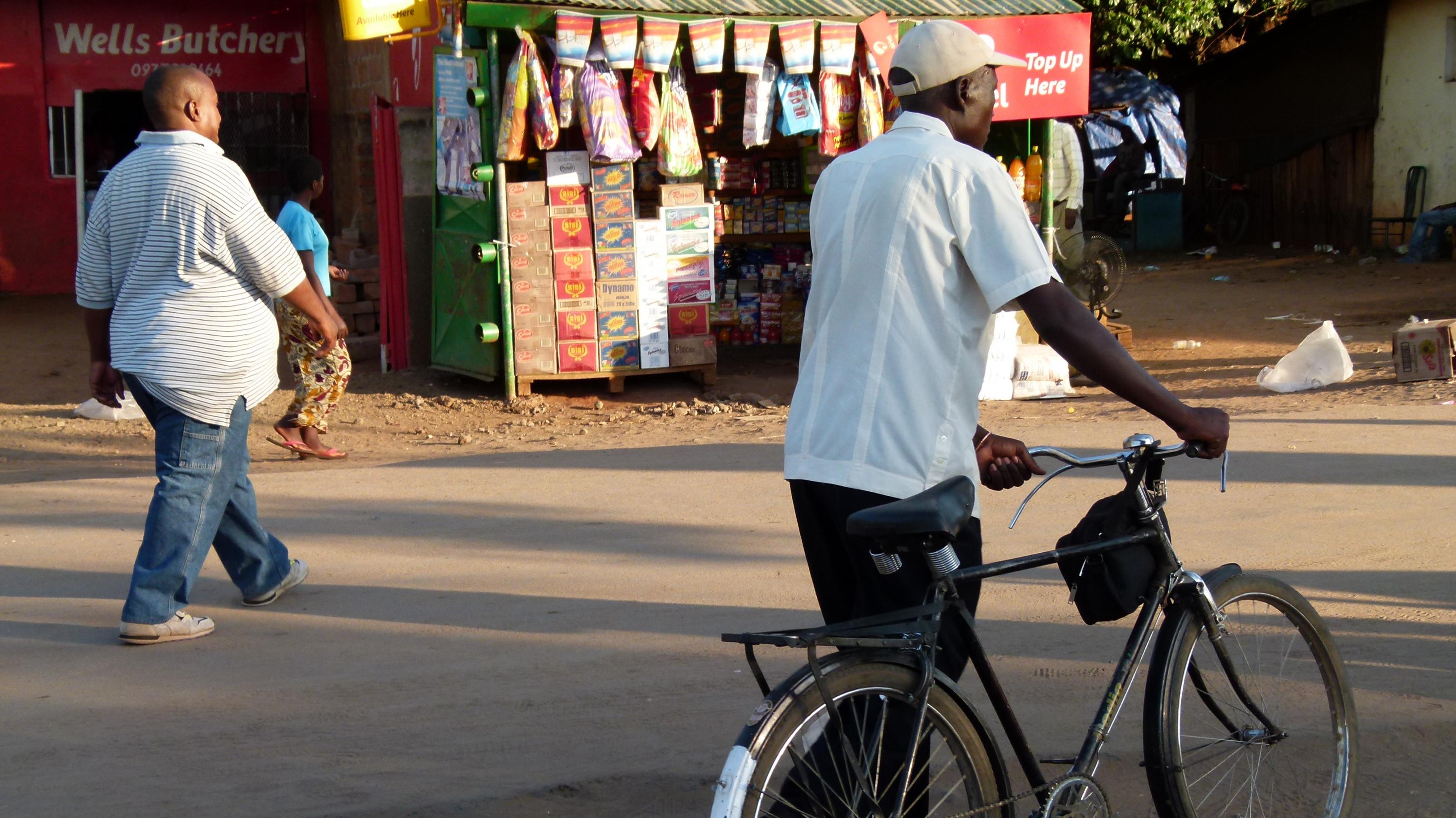 Straßenszene in Sambia: Zwei Fussgänger gehen an einem Markstand vorbei, einer schiebt sein Fahrrad