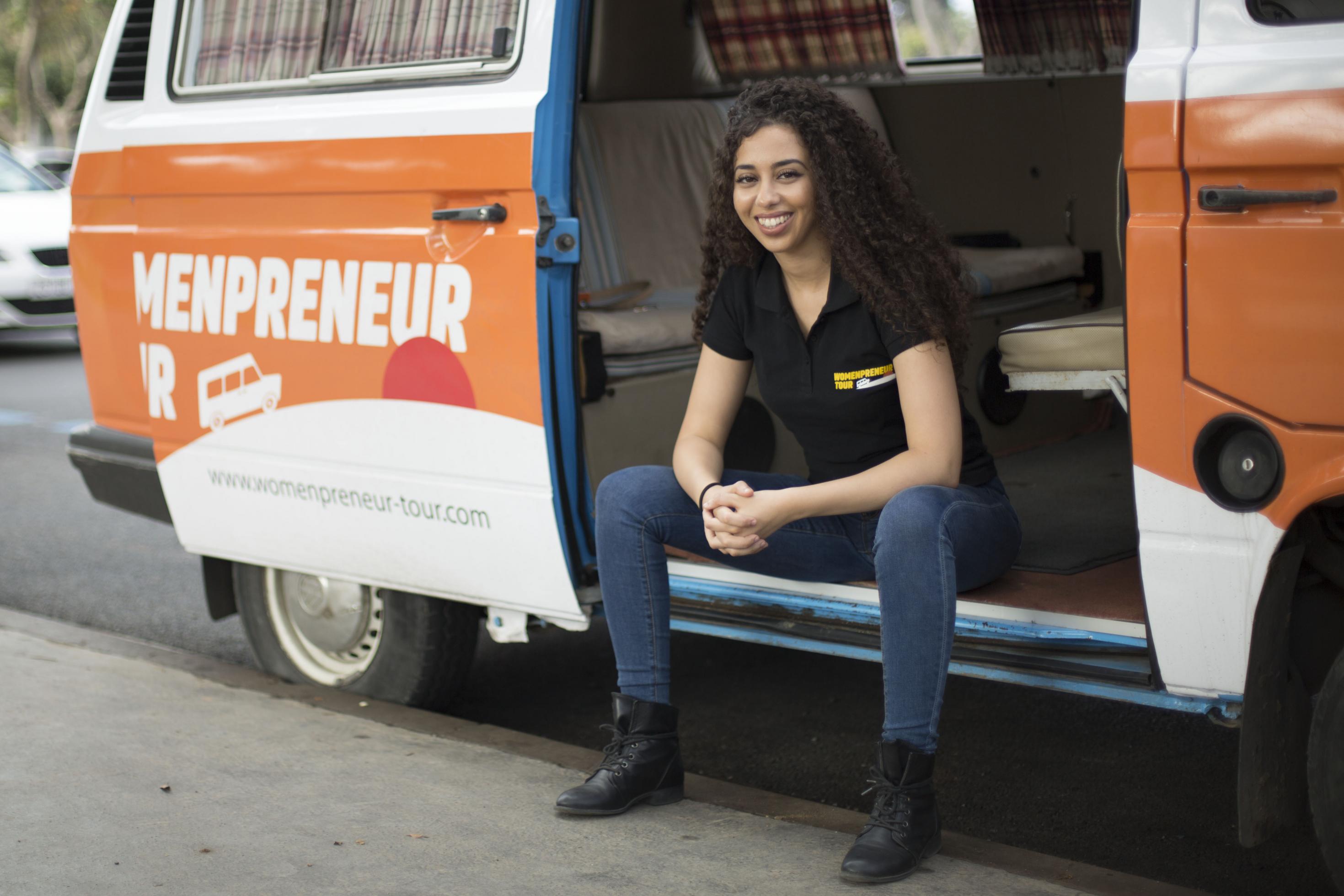 Eine junge Frau sitzt in der offenen Schiebetür eines Campingbusses, auf dem Bus und ihrem T-Shirt steht Womenpreneur Tour