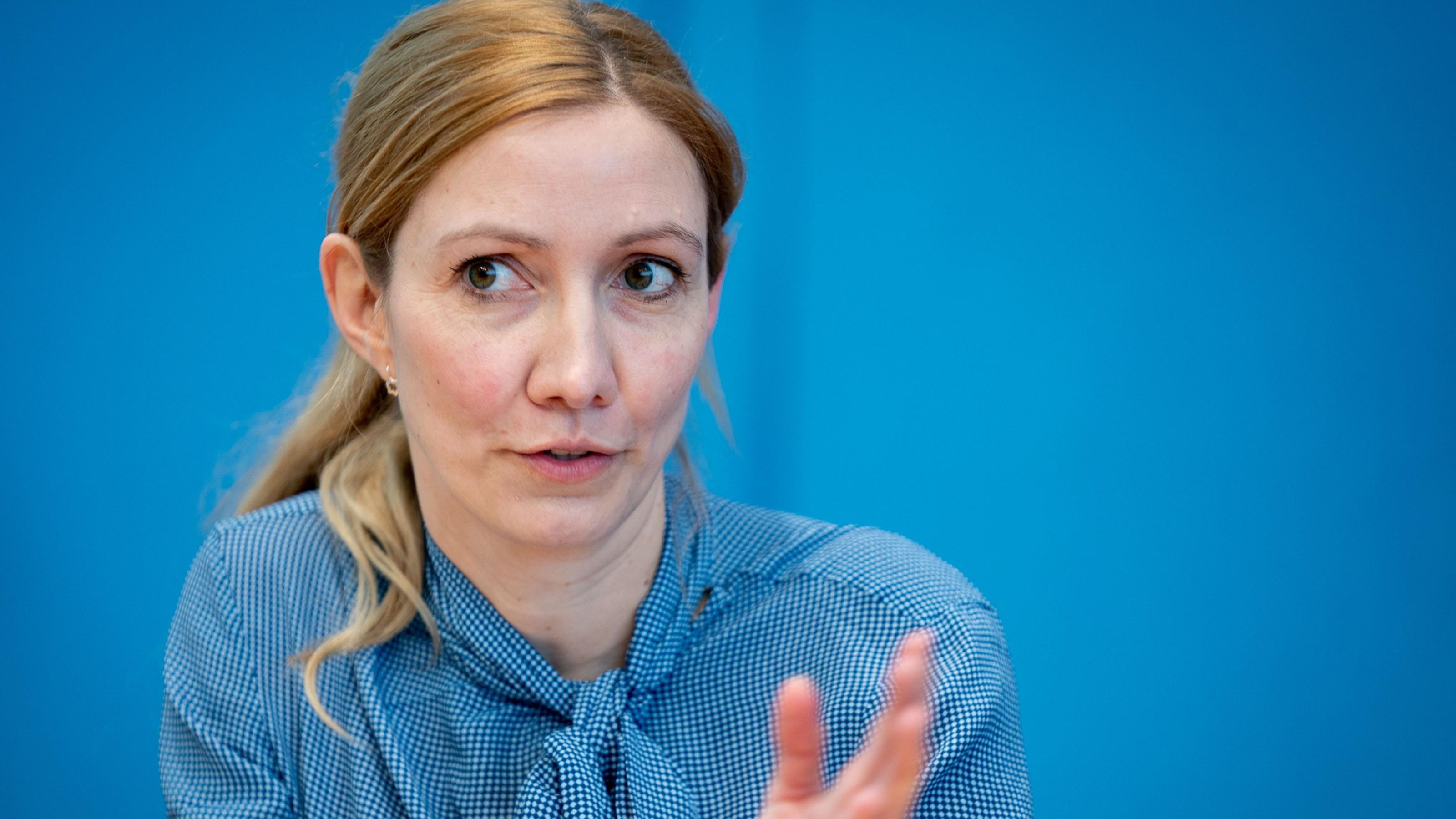 Eine Frau mit blonden Haaren und Pferdeschwanz sitzt gestikulierend vor eine blauen Wand.