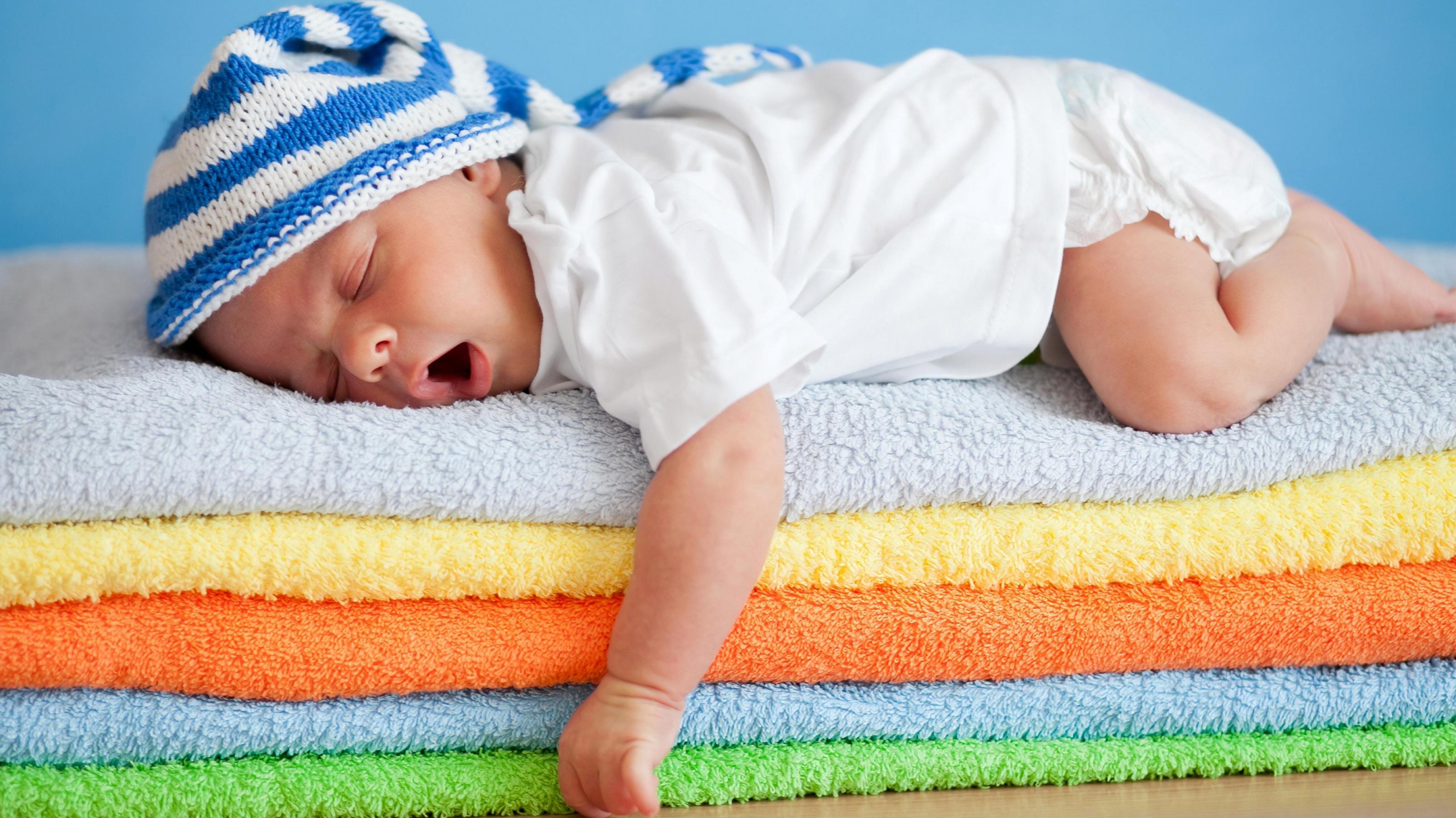 Ein Säugling mit einer blauweiß geringelten Mütze schläft vollkommen entspannt mit offenem Mund auf einem bunten Wäschestapel..