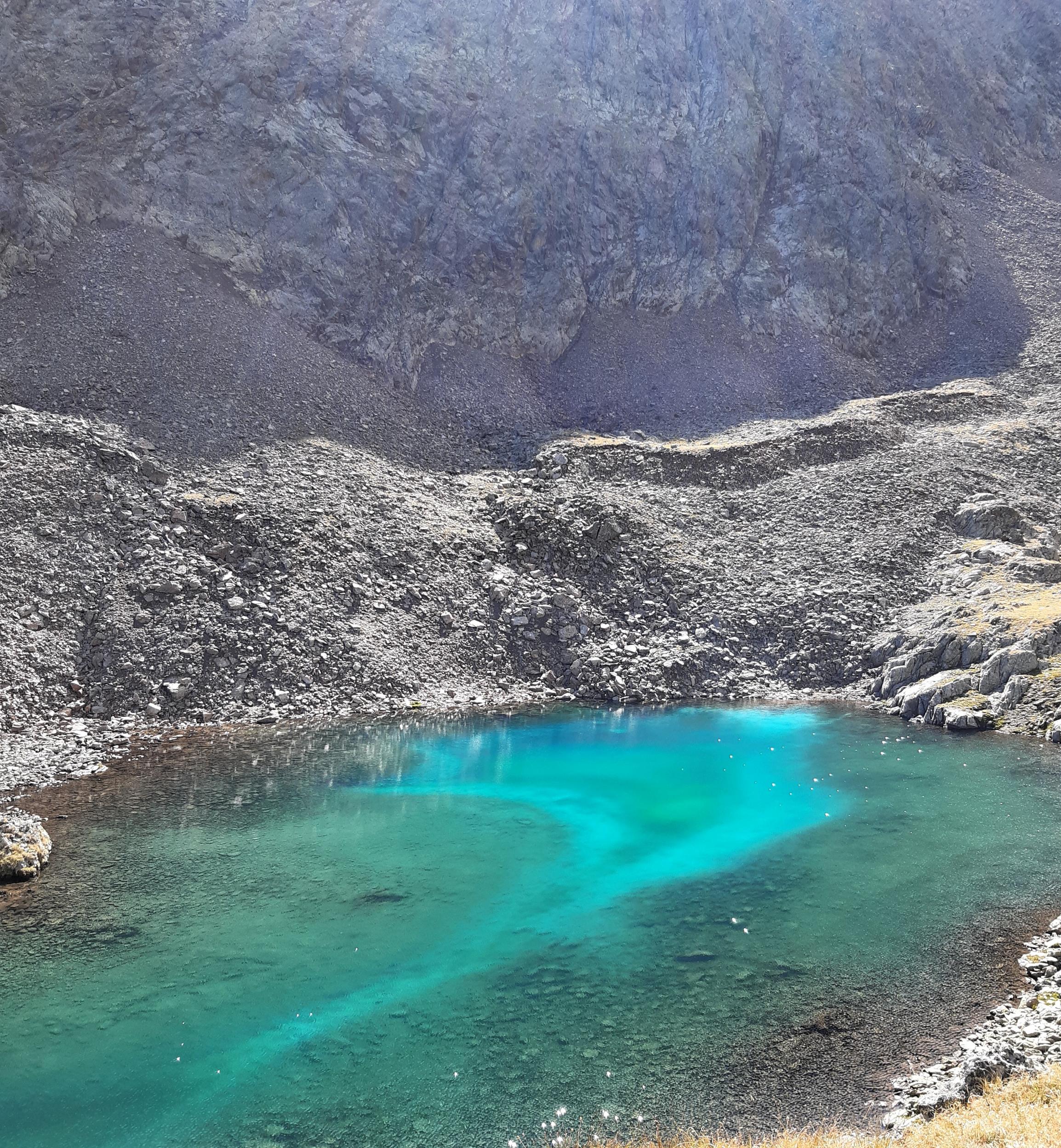 Vor einer dinklen Geröllhalde liegt transparent-grünes Wasser, in dem sich weiß-blaue Schlieren entlangziehen.