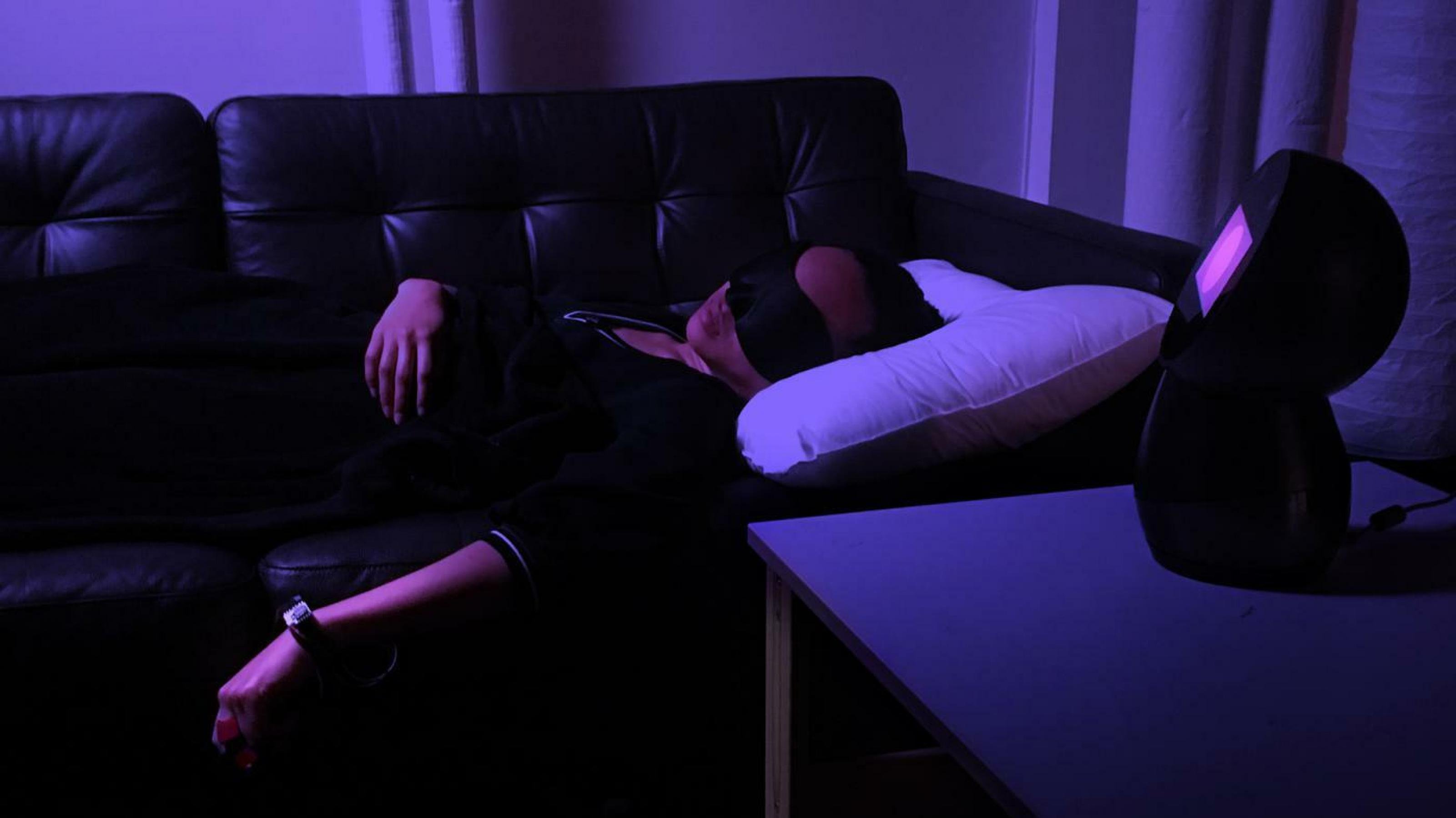 Eine Frau liegt in einem abgedunkelten Raum mit Schlafmaske auf einem Sofa. An den Fingern ihrer linken Hand sind kleine Sensoren angebracht.
