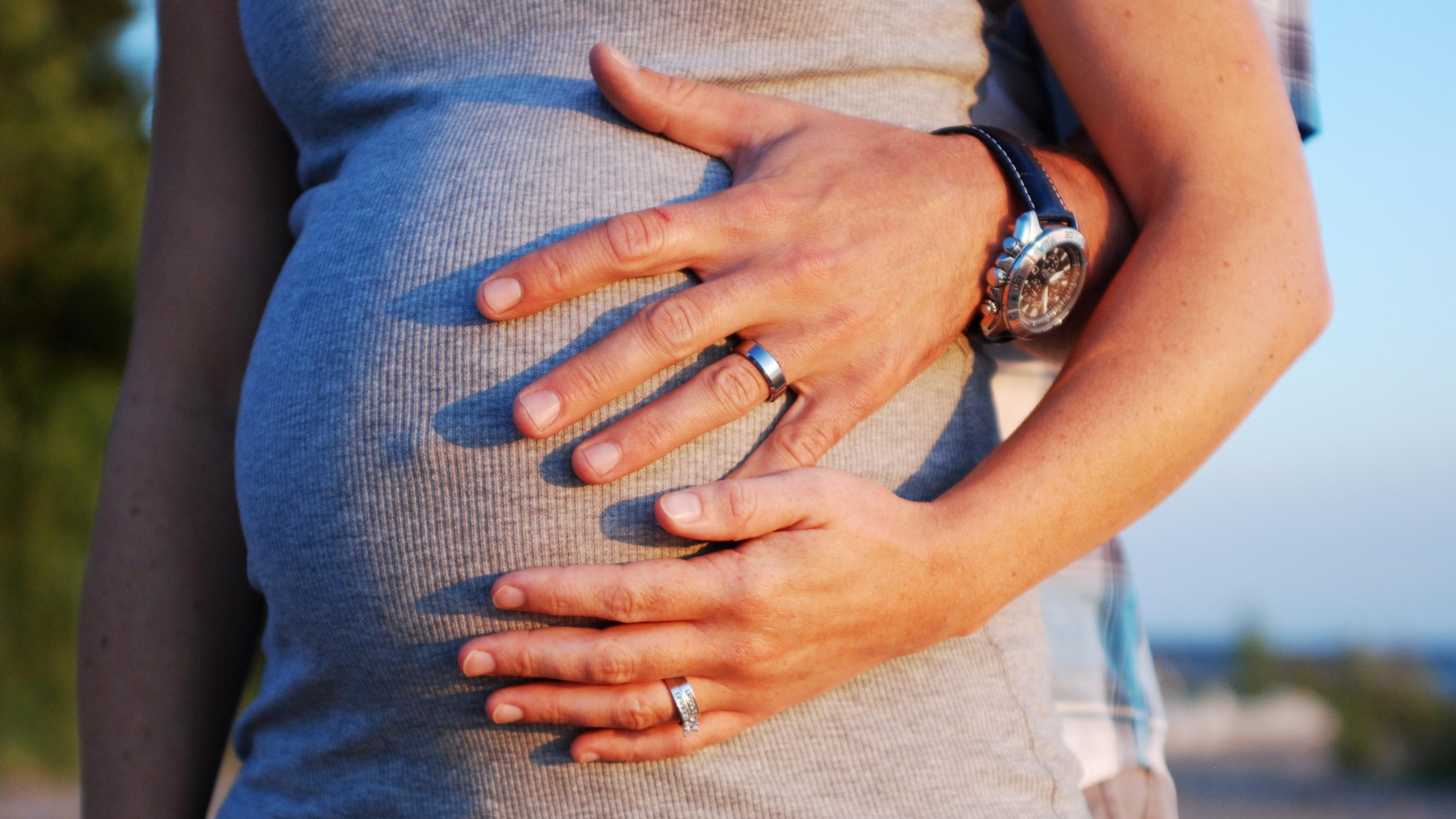 Auf dem Foto ist der Bauch einer schwangeren Frau zu sehen. Die Frau selbst und eine andere Person, die hinter der Frau steht, halten ihre Hand auf den Bauch.