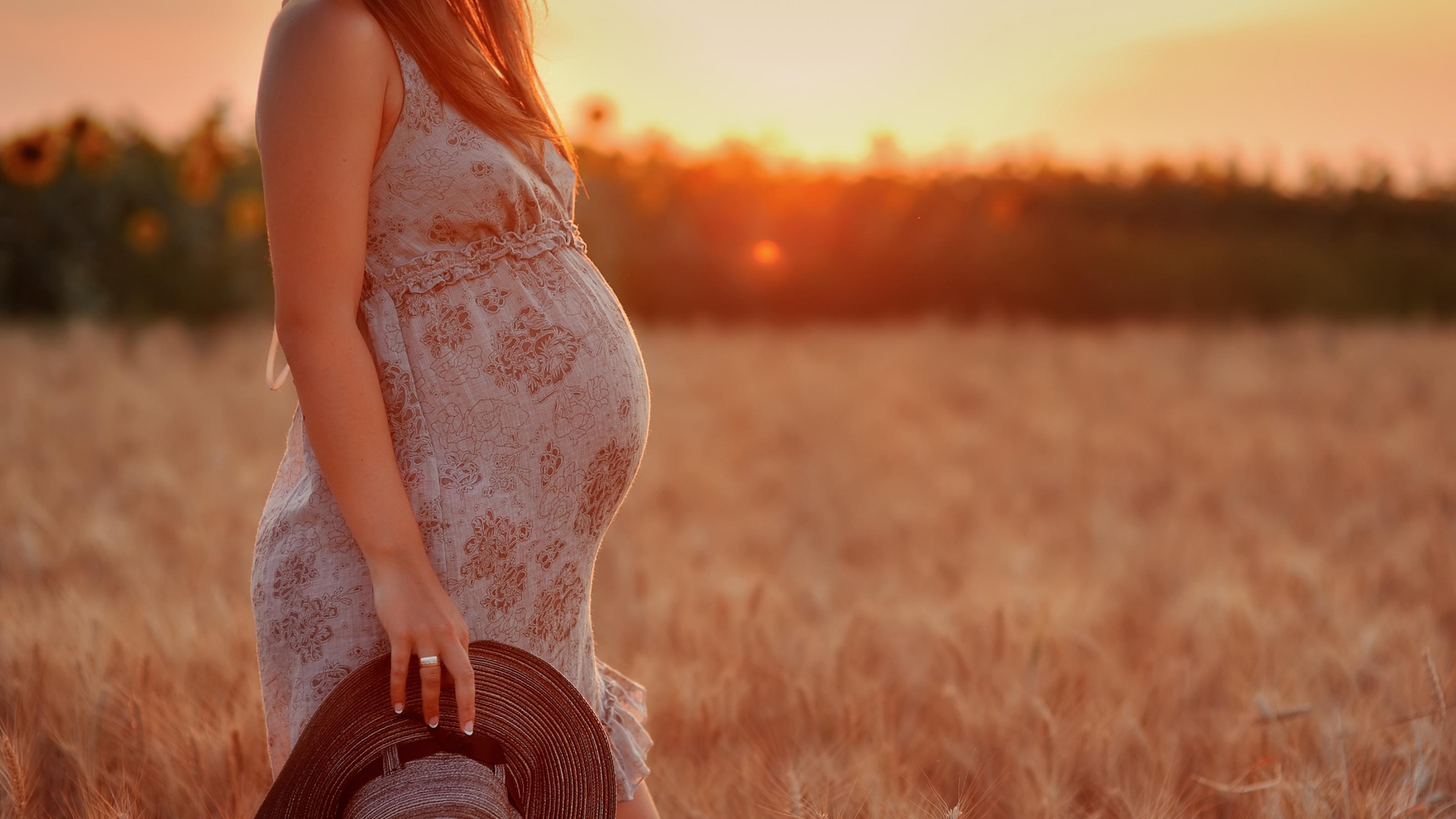 Vor einem Weizenfeld bei Sonnenuntergang ist eine hochschwangere Frau zu sehen. Sie trägt ein leichtes Sommerkleid und hält in der Hand des herabhängenden Arms einen Sonnenhut.