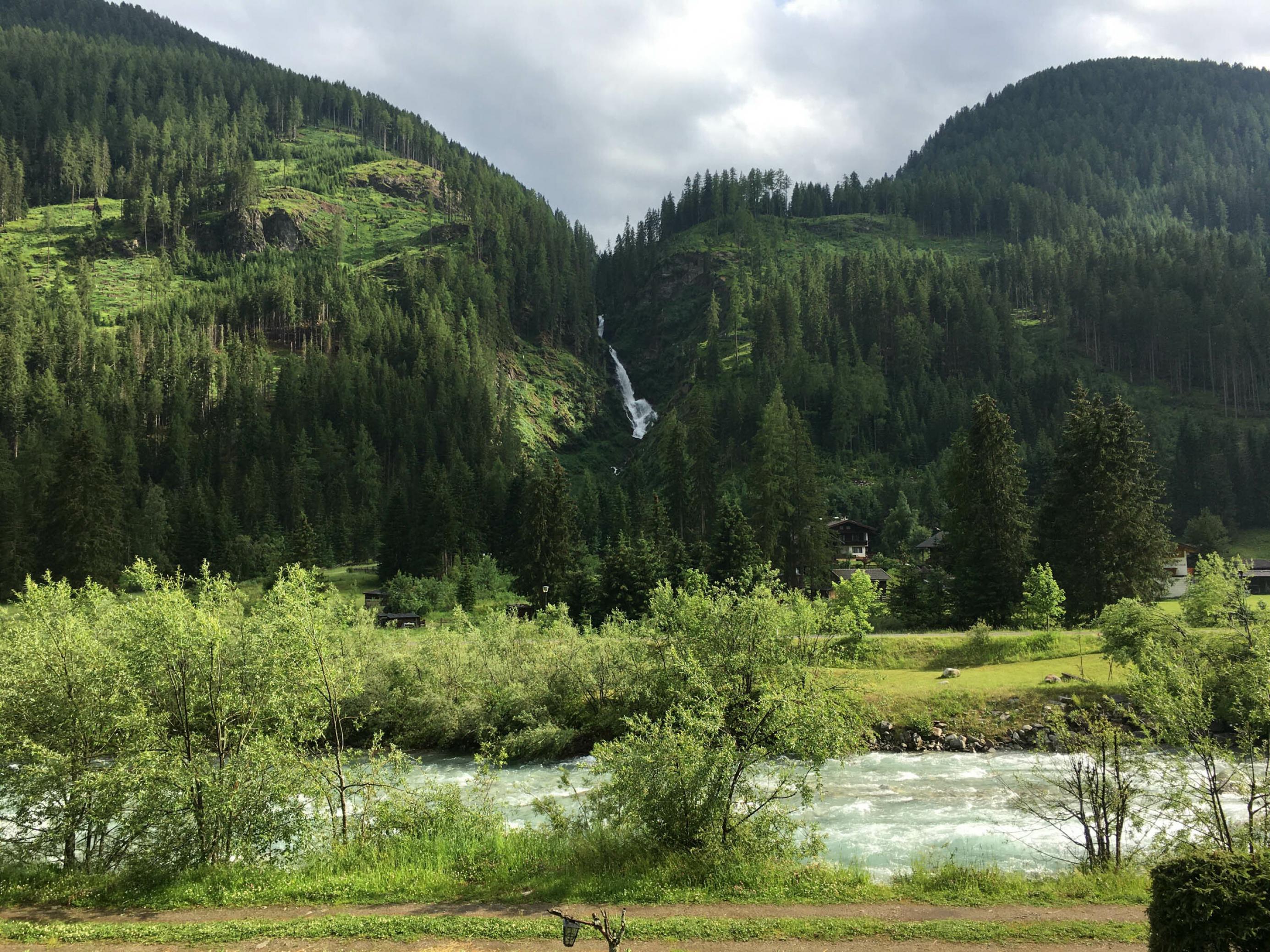 Quer im Bild ein Gebirgsbach mit Uferghölz, geradeaus Berge, Wald und ein hoher Wasserfall.