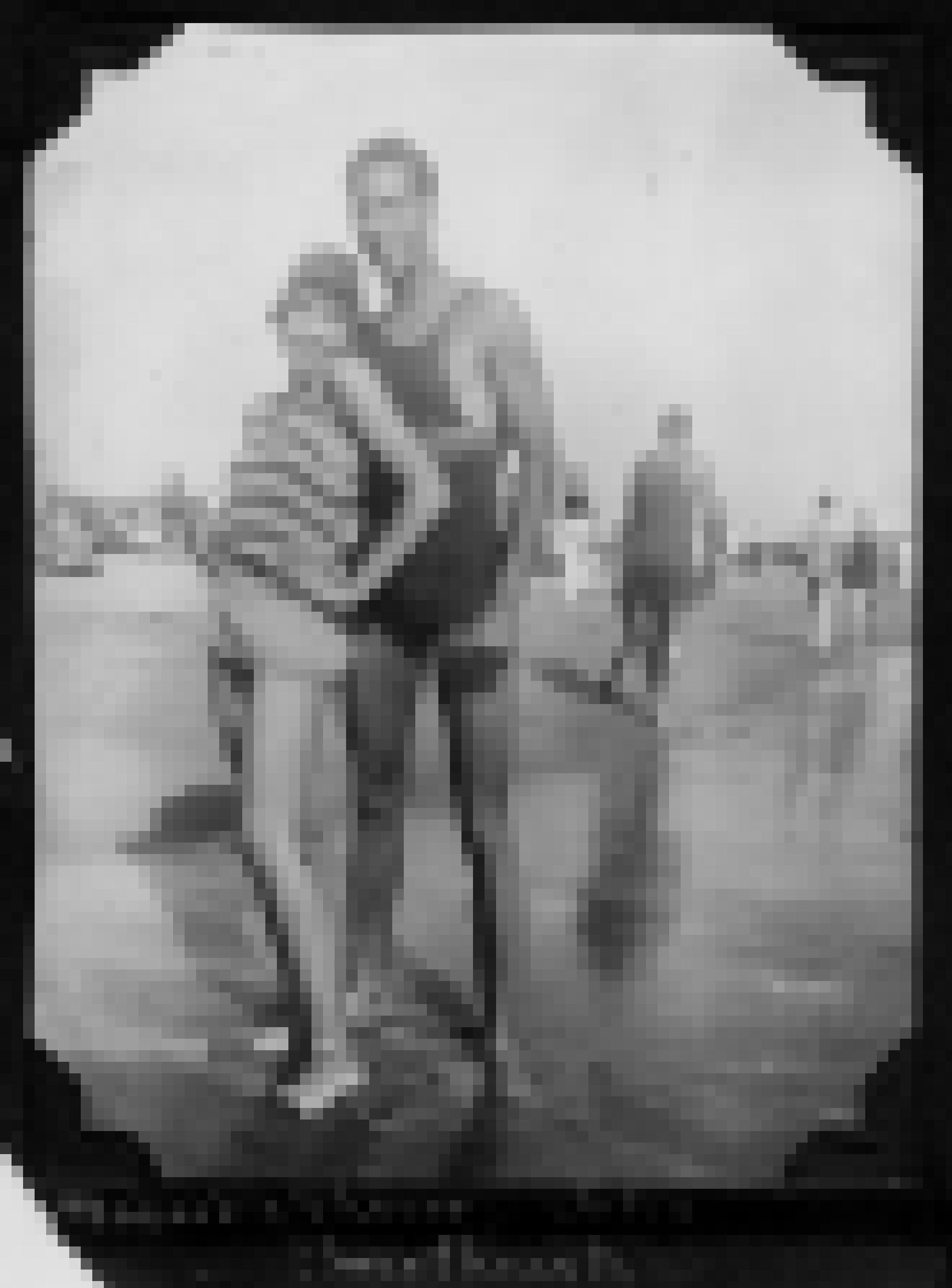 Schwarz-Weiß Foto von einem jungen Paar in Badekleidung der 20er Jahre. Sie posieren am Strand für die Kamera. Hinter ihnen Flaneure, Badende und Sonnenbadende.