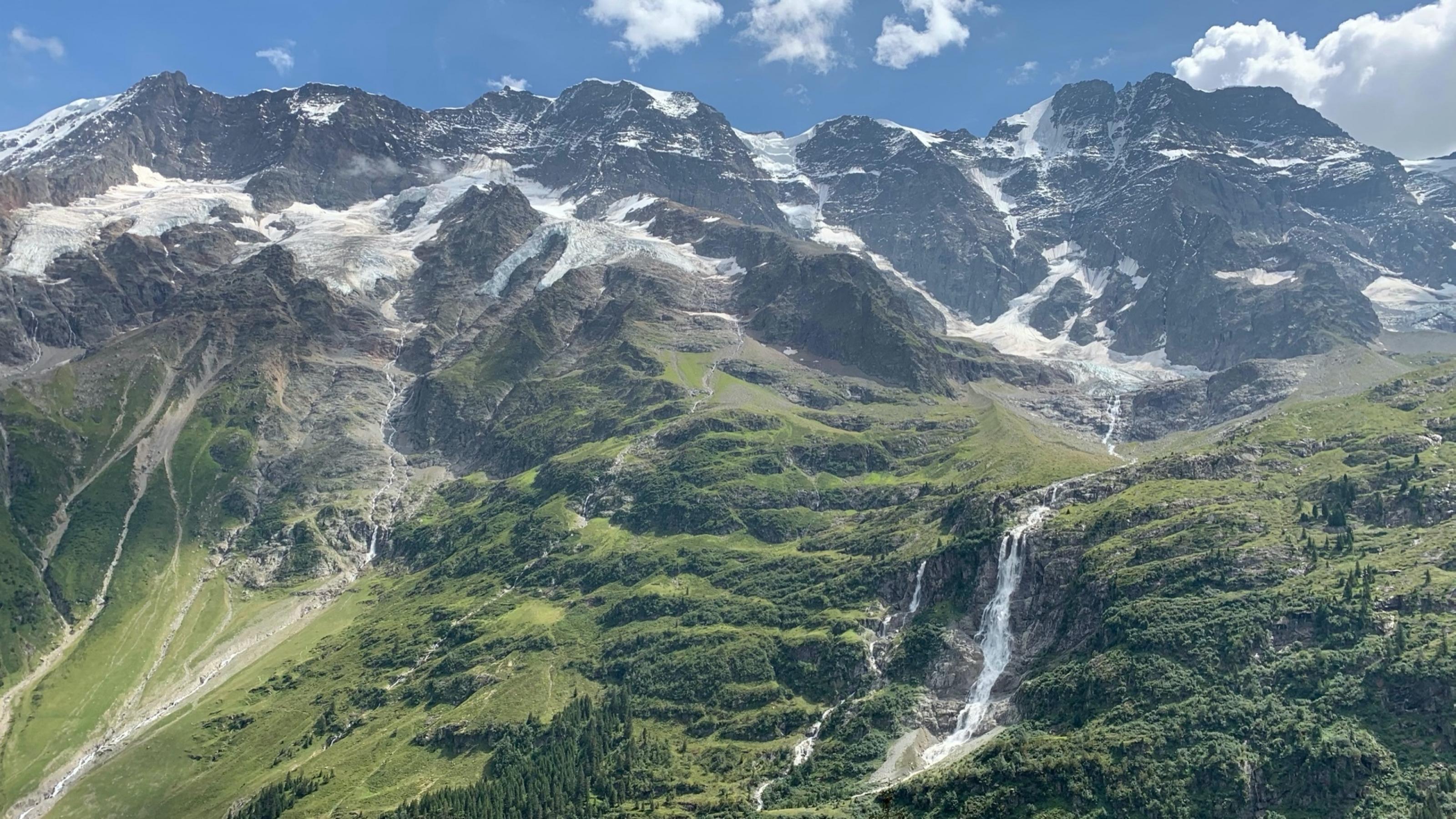 Schneebedeckte Berge ragen in den blauen Himmel. Die Vegetation verändert sich talabwärts von Gestein zu Wald- und Grünflächen. Mittig befindet sich ein Wasserfall.